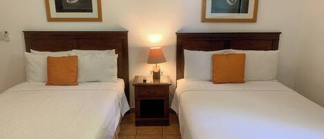 Two single beds with white linens and orange pillows, a nightstand with a lamp, and framed artwork on the walls.