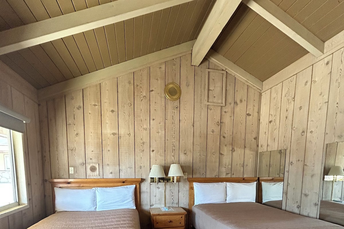 A room with two beds, wooden walls, a wooden ceiling, and a small wooden nightstand with lamps.