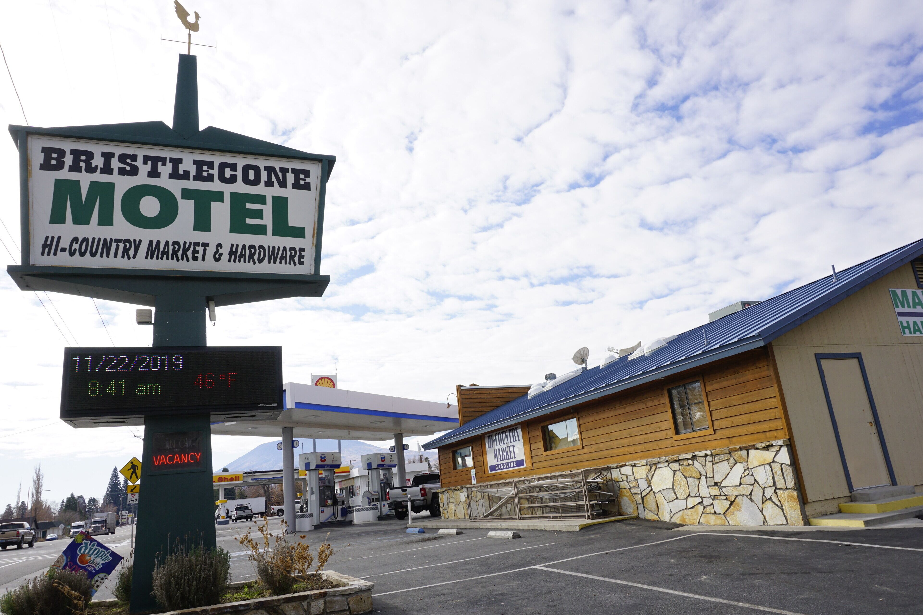 A motel sign with a date and temperature display, a gas station in the background, and a wooden building with a sign for a market and hardware store.