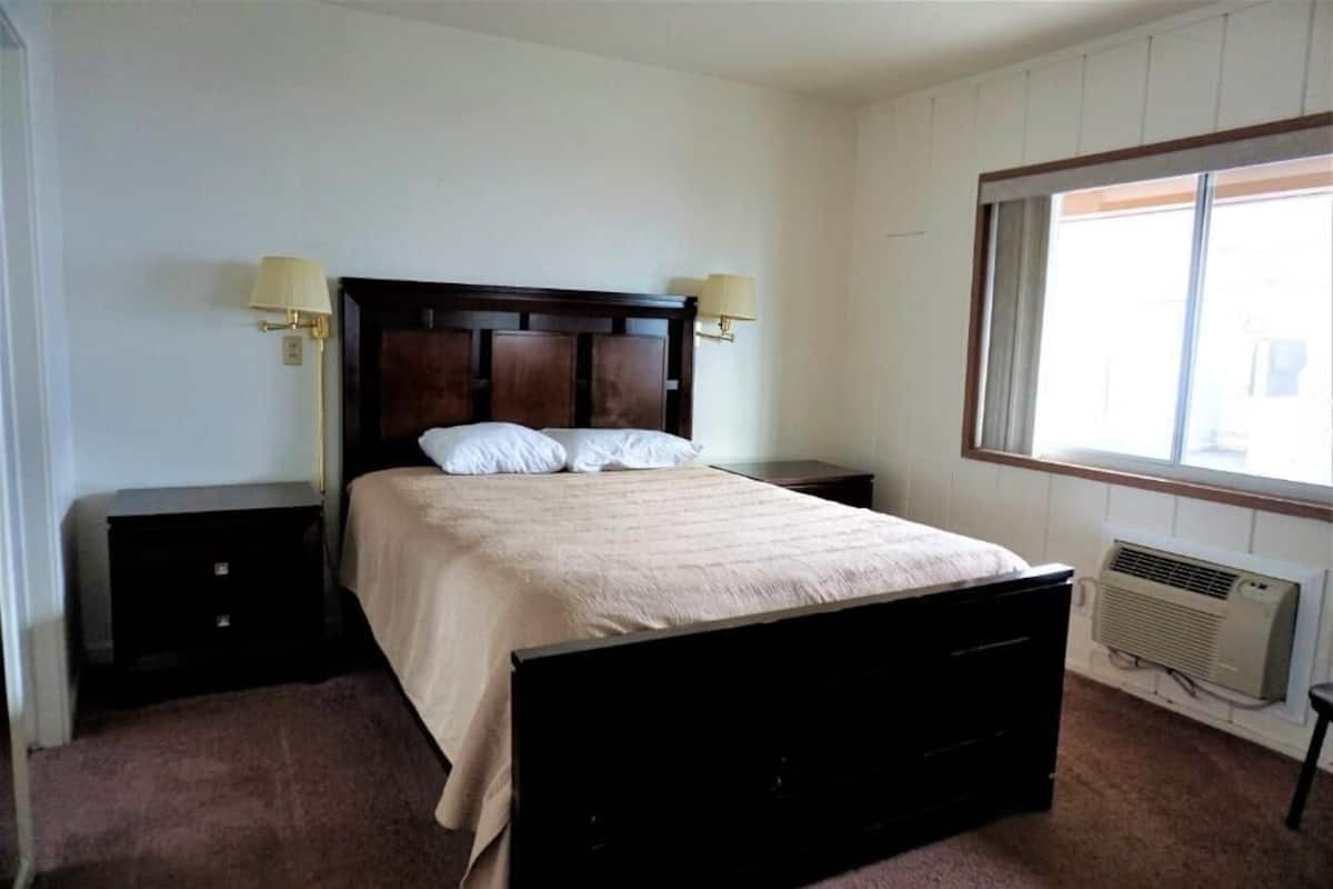 A bedroom with a dark wooden bed, two bedside tables with lamps, a window with blinds, and a wall-mounted air conditioner.
