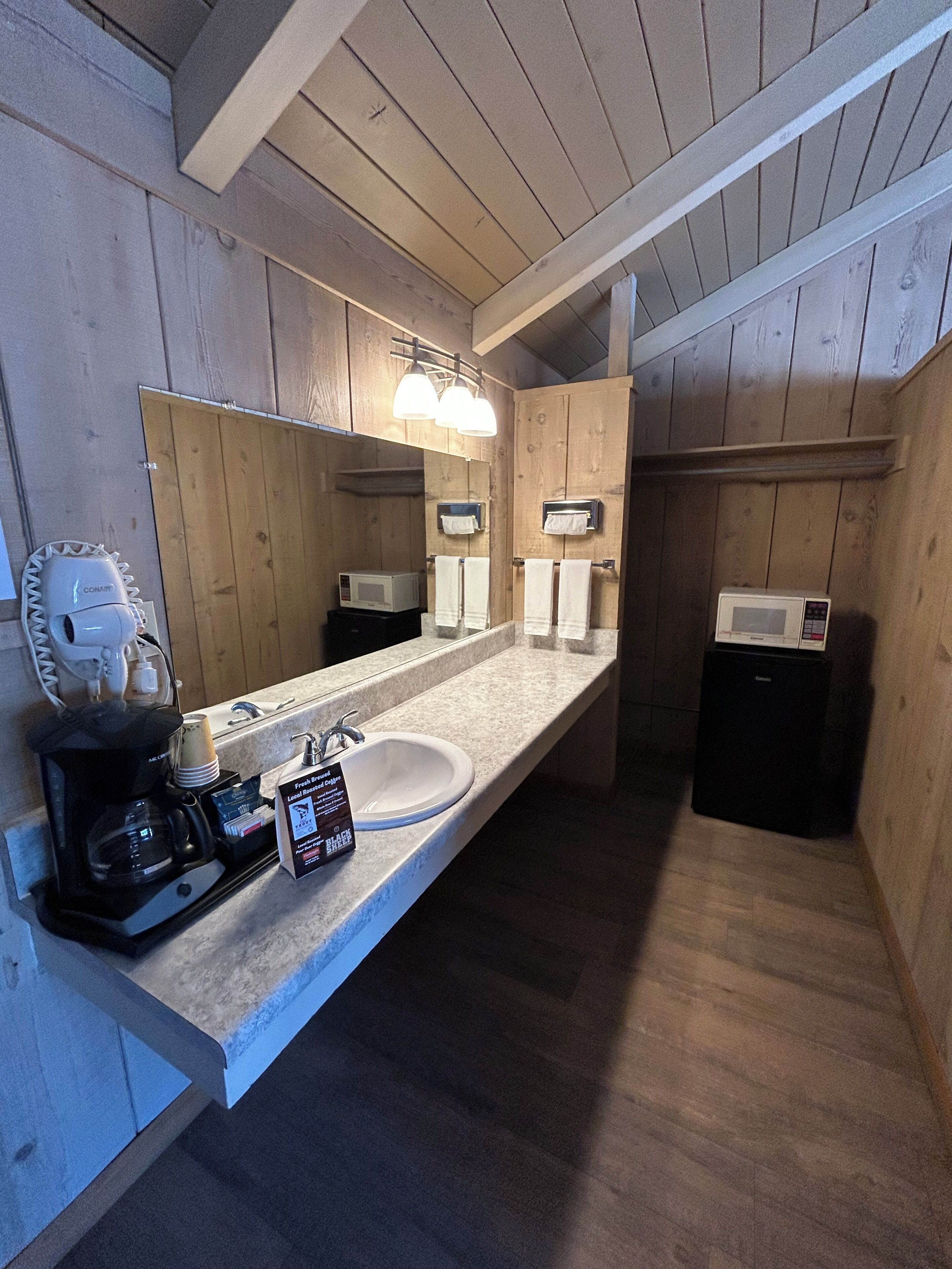 Une salle de bain de style rustique, avec des murs en bois, un grand miroir, deux distributeurs de serviettes, une machine à café et un four à micro-ondes.