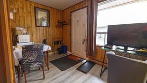 Habitación pequeña con mesa de comedor, dos sillas, televisor sobre una mesa de vidrio y una puerta con perchero.