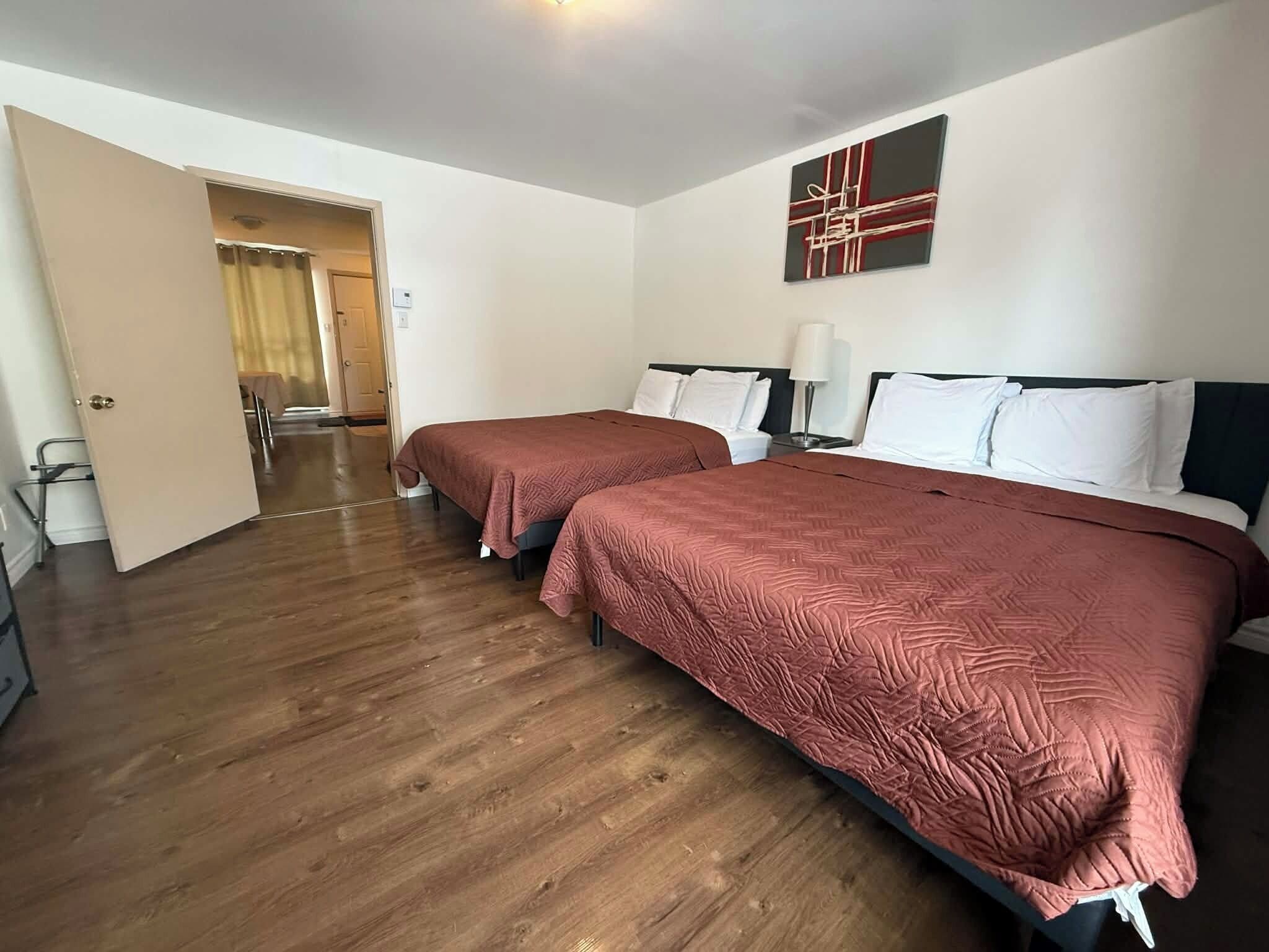 A hotel room with two beds, wooden flooring, a door leading to a hallway, and a painting on the wall.