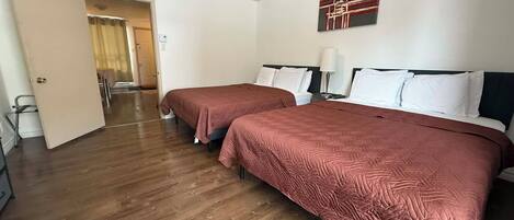 A hotel room with two beds, wooden flooring, a door leading to a hallway, and a painting on the wall.