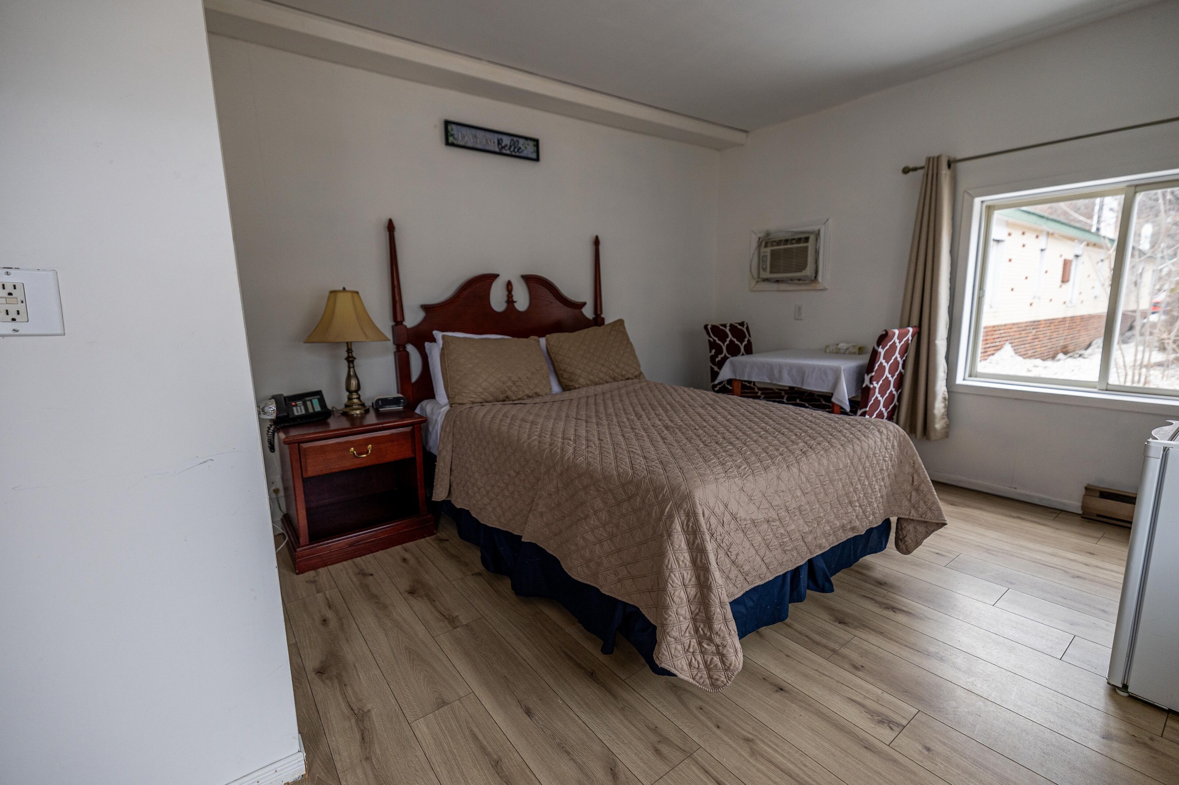 Un dormitorio con una cama de madera, una mesita de noche con lámpara, una pequeña mesa con una silla y una ventana con cortinas.