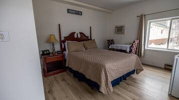 A bedroom with a wooden bed, a nightstand with a lamp, a small table with a chair, and a window with curtains.