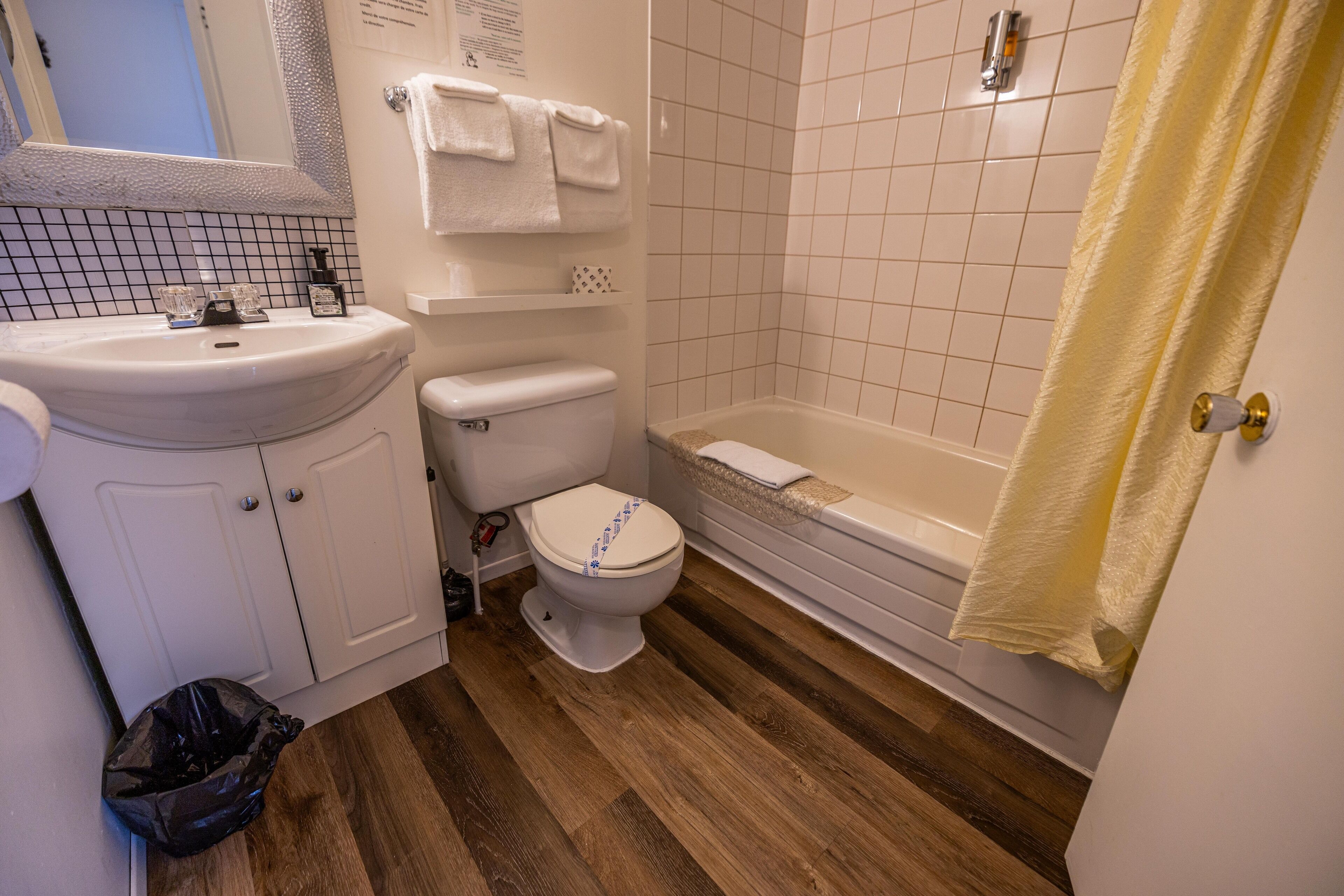 A bathroom with a white sink, toilet, and bathtub. There is a black trash bag on the floor.