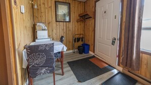 Habitación pequeña con pared de madera, puerta blanca, una mesa con mantel, una silla, un pequeño calentador, perchero y un cubo de basura azul.