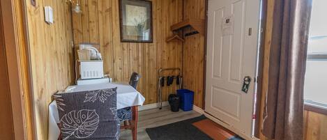 Une pièce de taille modeste avec un mur en bois, une porte blanche, une table recouverte d’une nappe, une chaise, un petit radiateur, un porte-manteau et une poubelle bleue.