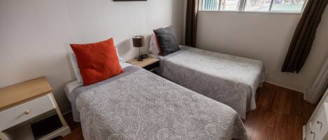 A small, tidy room with two single beds, a nightstand, a lamp, and a window with curtains.