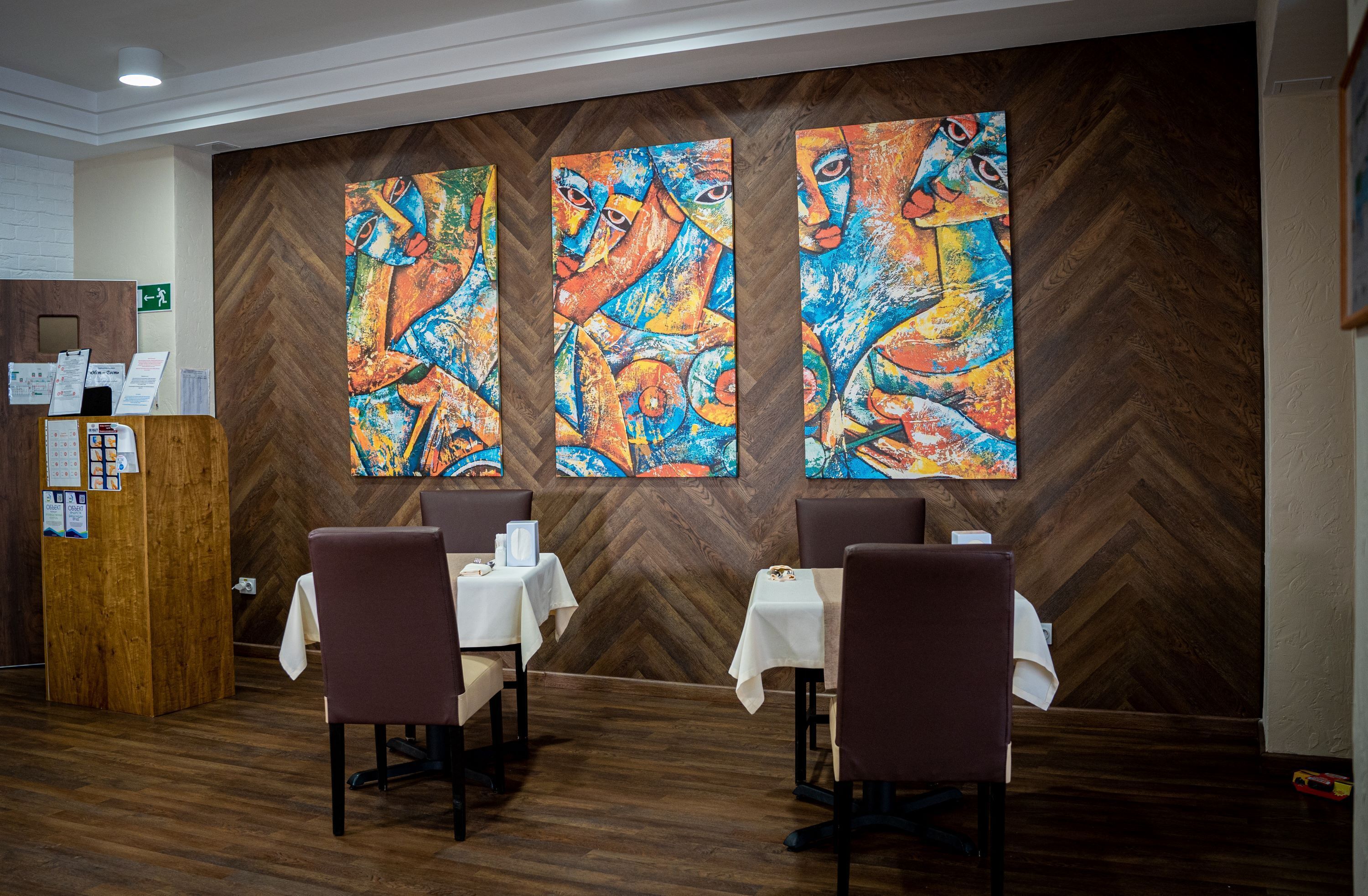 Een restaurant met houten visgraatvloer, witte tafels met witte tafellinnen, en abstracte schilderijen aan de muur.