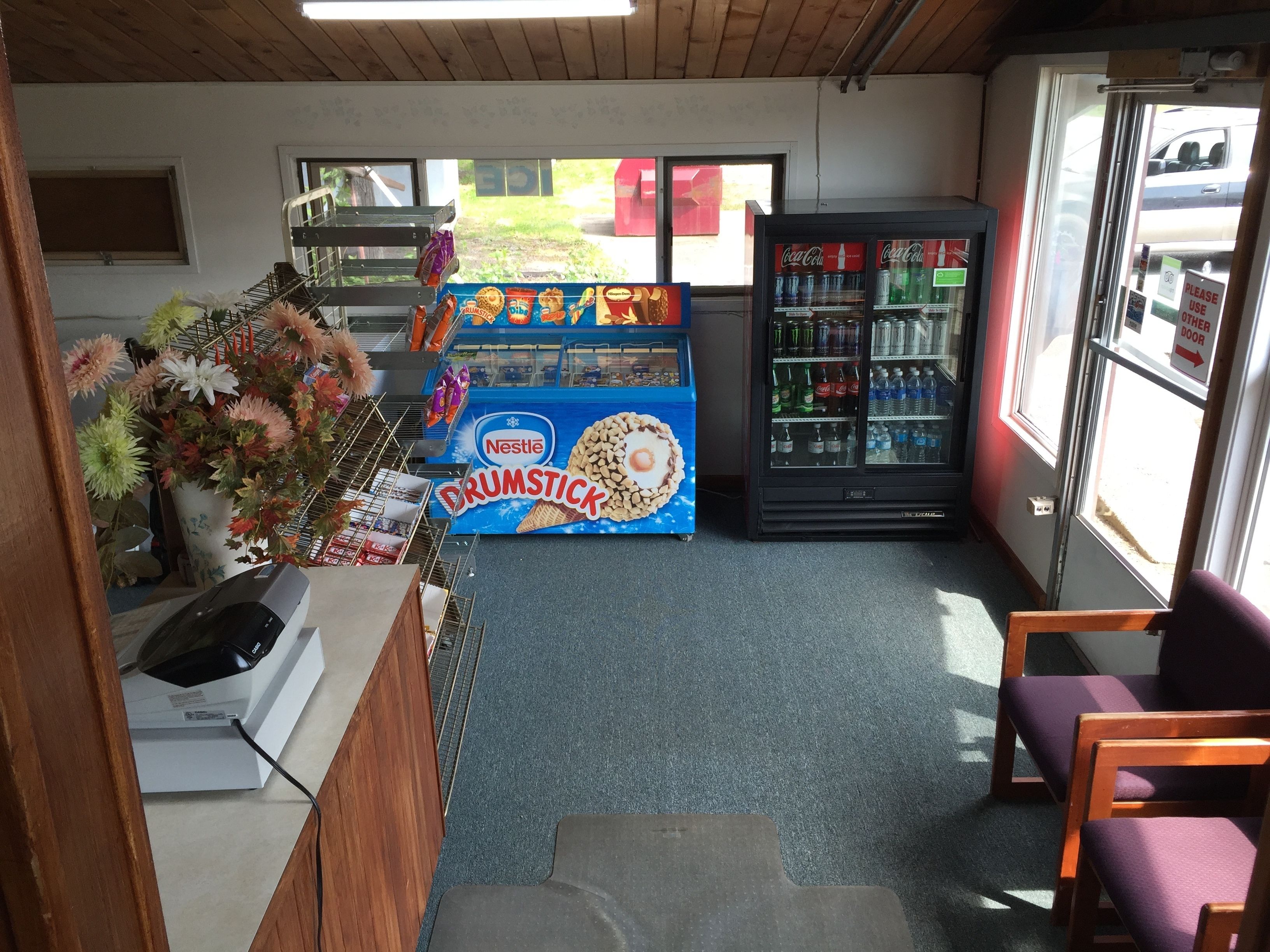 Una pequeña tienda de conveniencia con una exhibición de aperitivos y bebidas, una máquina expendedora y una silla.