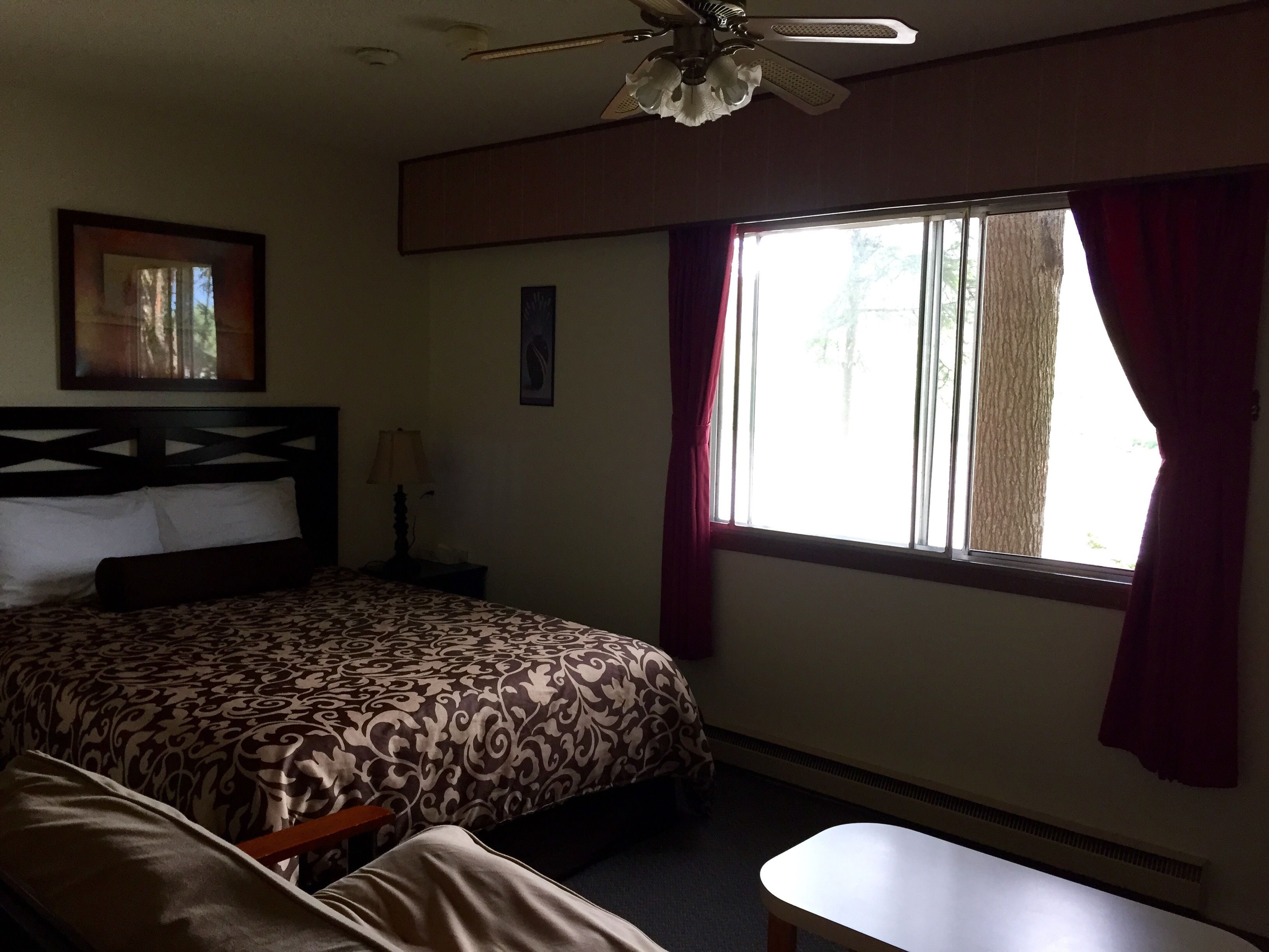 A hotel room with a bed, a desk, a ceiling fan, a window with curtains, and a framed picture on the wall.