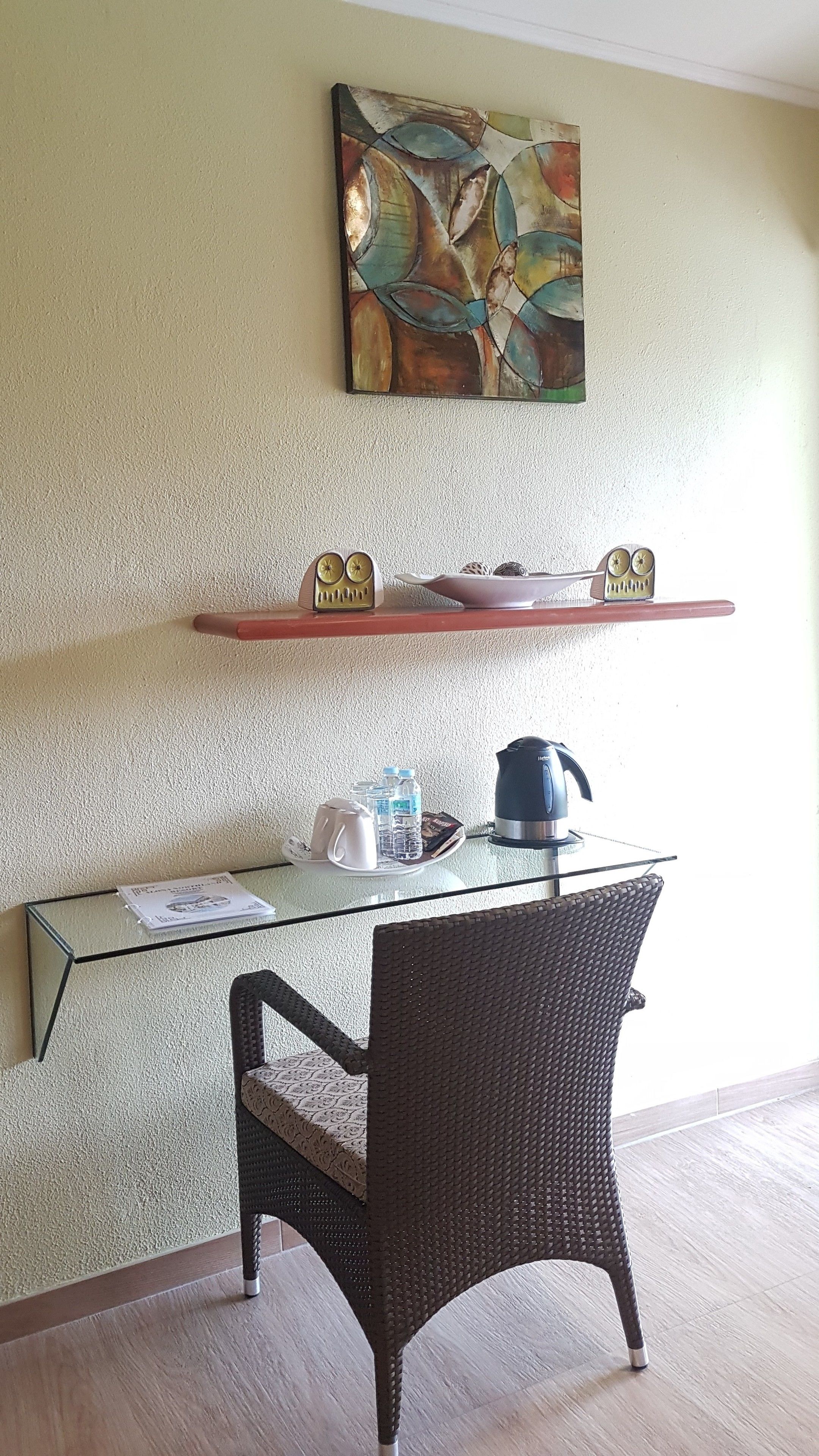 A dining area with a wicker chair, a glass table, a kettle, and a shelf with decorative items.