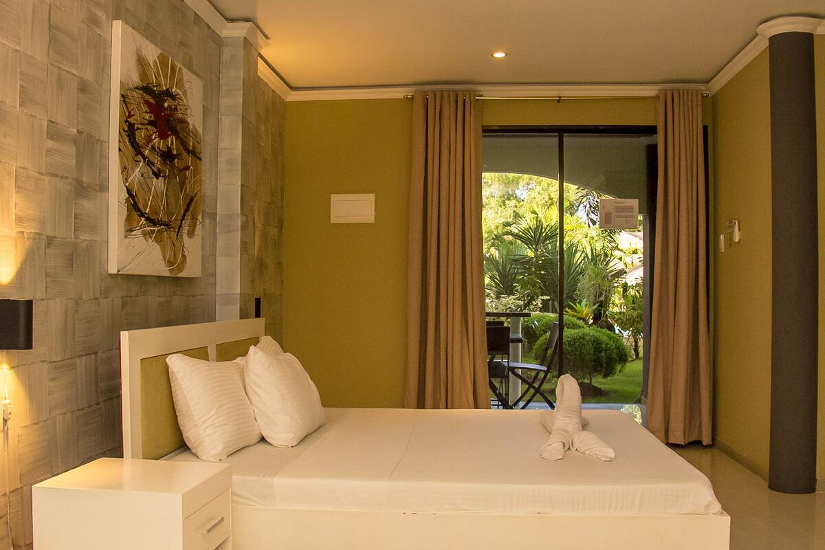 A hotel room with a large bed, a nightstand, a wall-mounted artwork, and a view of a garden through a sliding door.