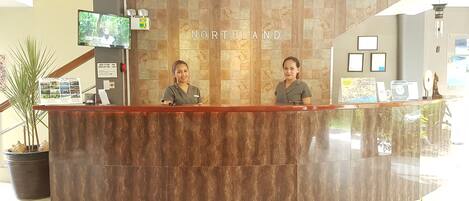 A reception desk with two staff members, a mounted television, and a welcome sign.