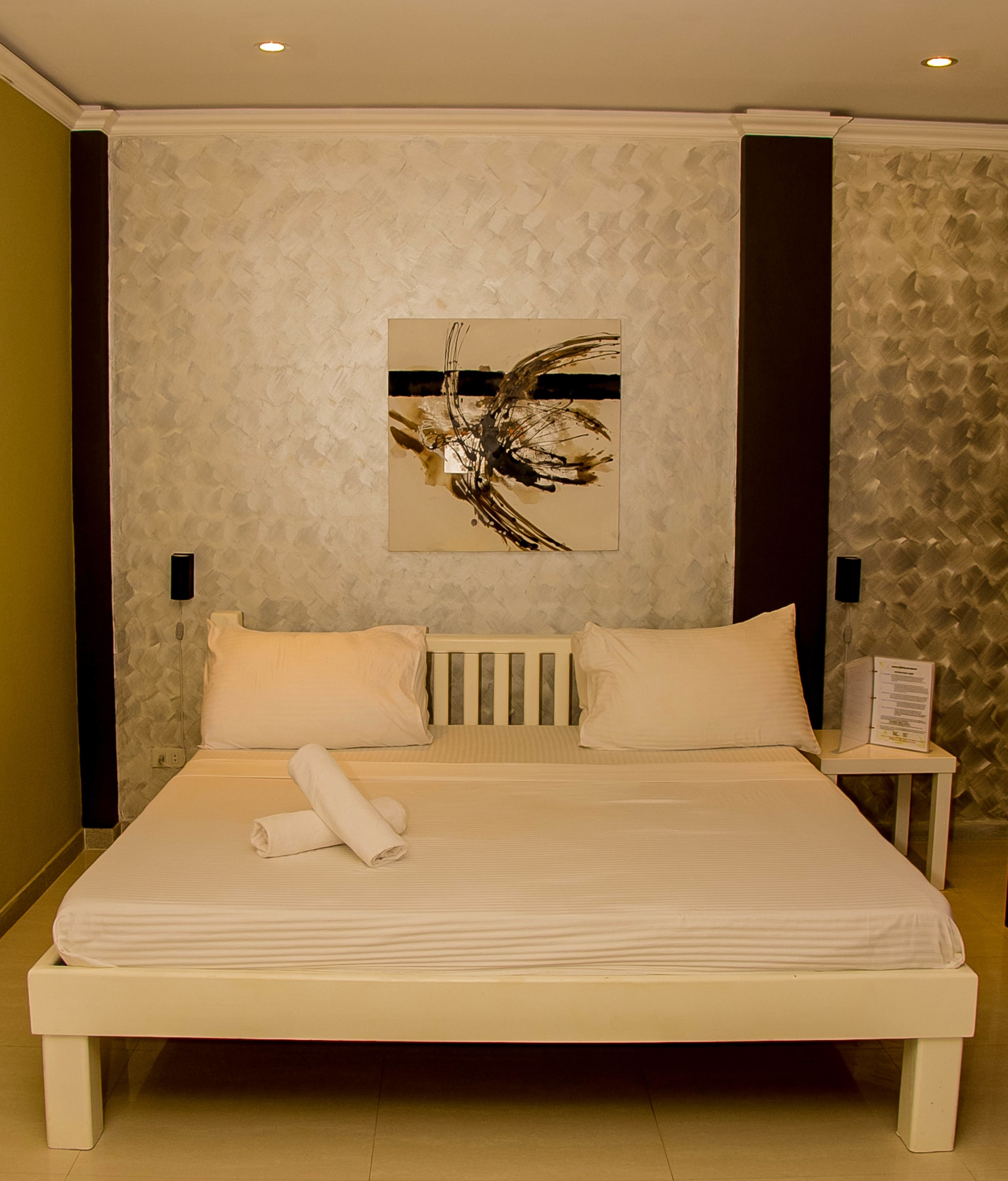 A neatly made bed with white linens and a rolled towel, a wall-mounted abstract painting, and a small bedside table with a book or menu.