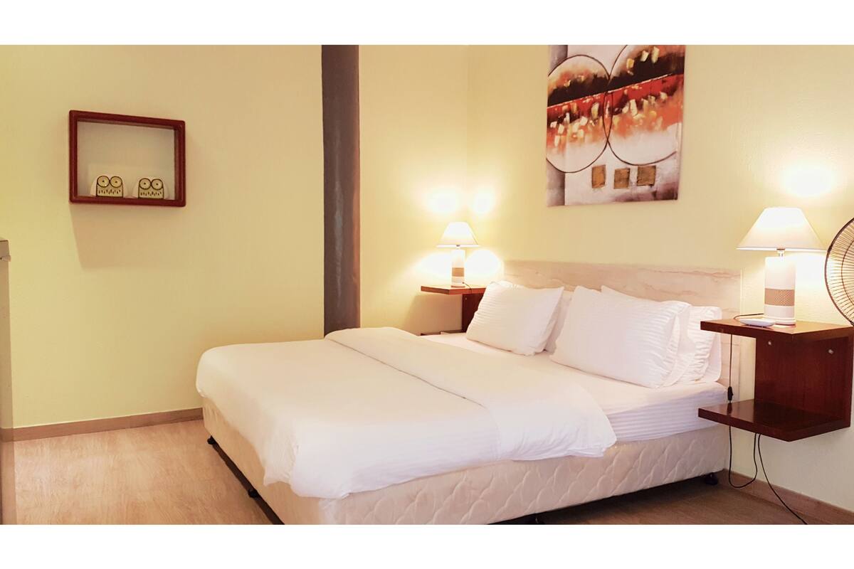 A neatly made bed with white linens, two bedside tables with lamps, a wall-mounted mirror, and a painting above the bed.