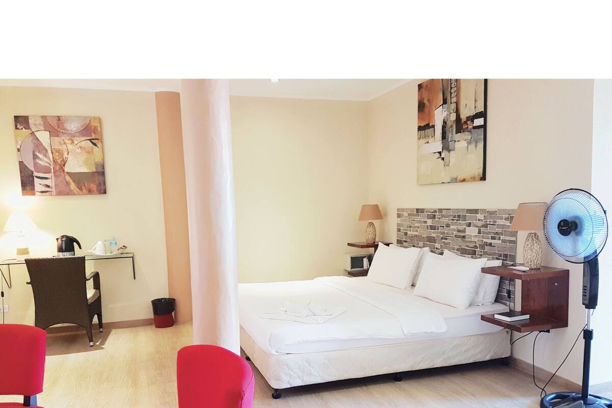 A hotel room with a bed, a desk, a chair, a standing fan, and artwork on the walls.