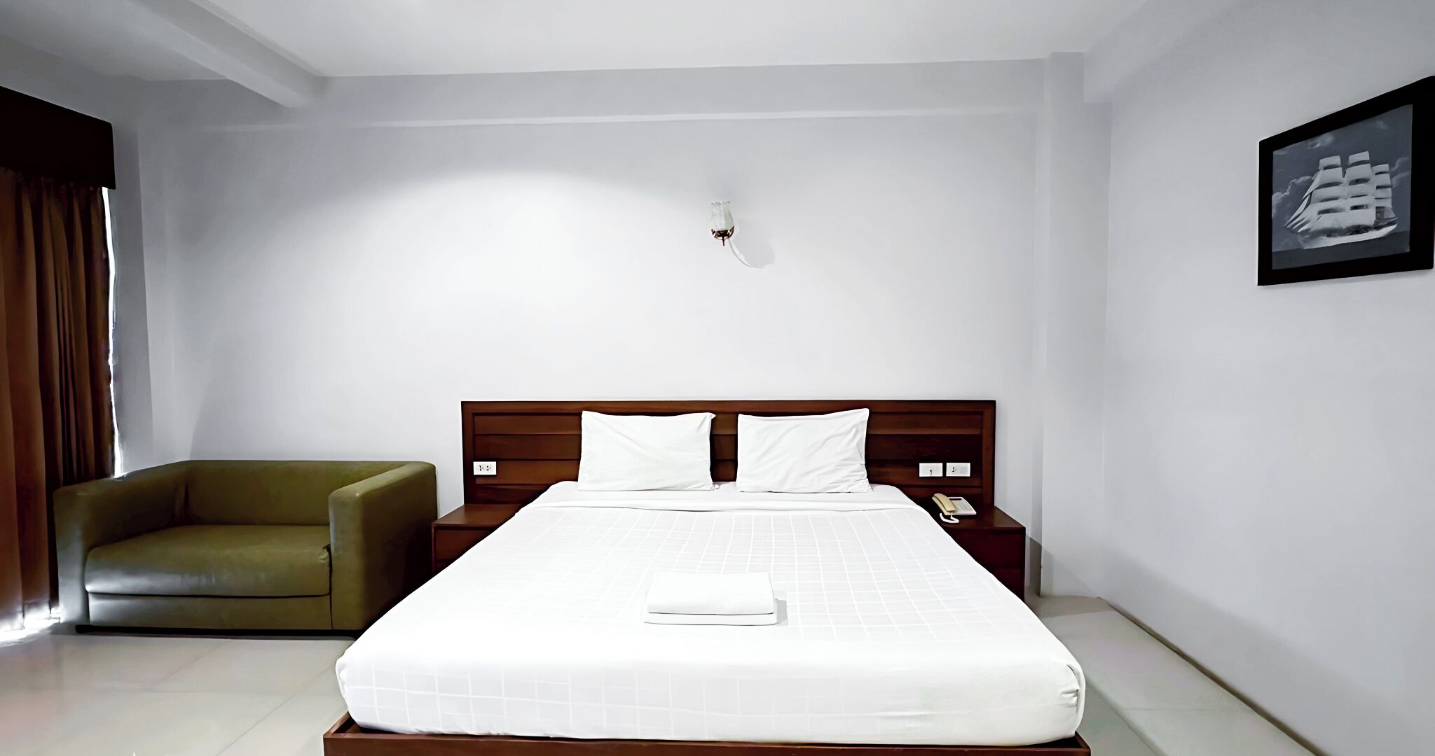 Una habitación de hotel con una cama grande, cabecero de madera, un sillón verde, una mesita de noche con un teléfono y un cuadro en la pared.