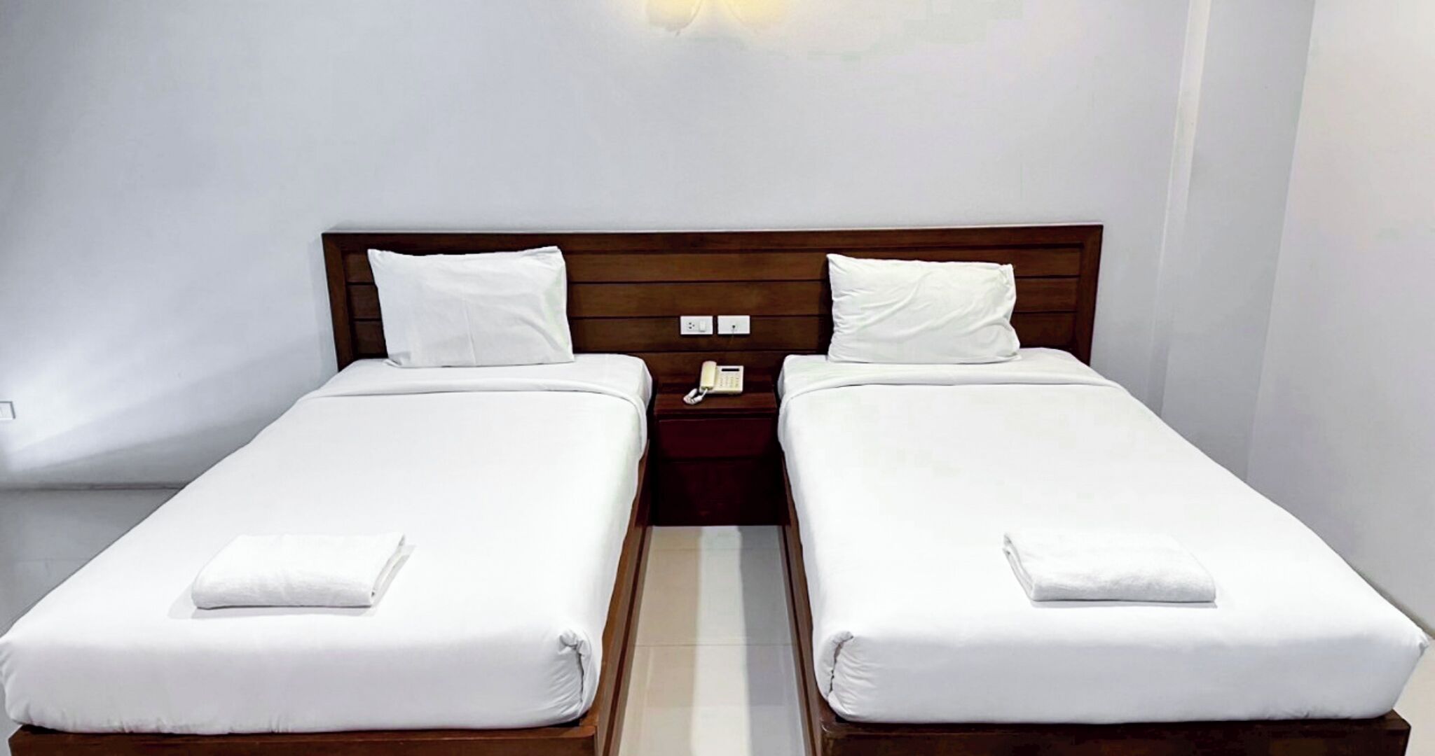 Cama doble con ropa blanca y cabecera de madera. Se ve una mesita de noche con un teléfono y una lámpara pequeña.