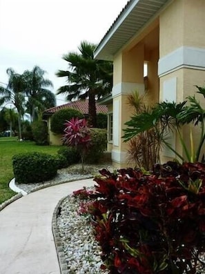 A building with a beige facade, a curved pathway, and a variety of tropical plants.