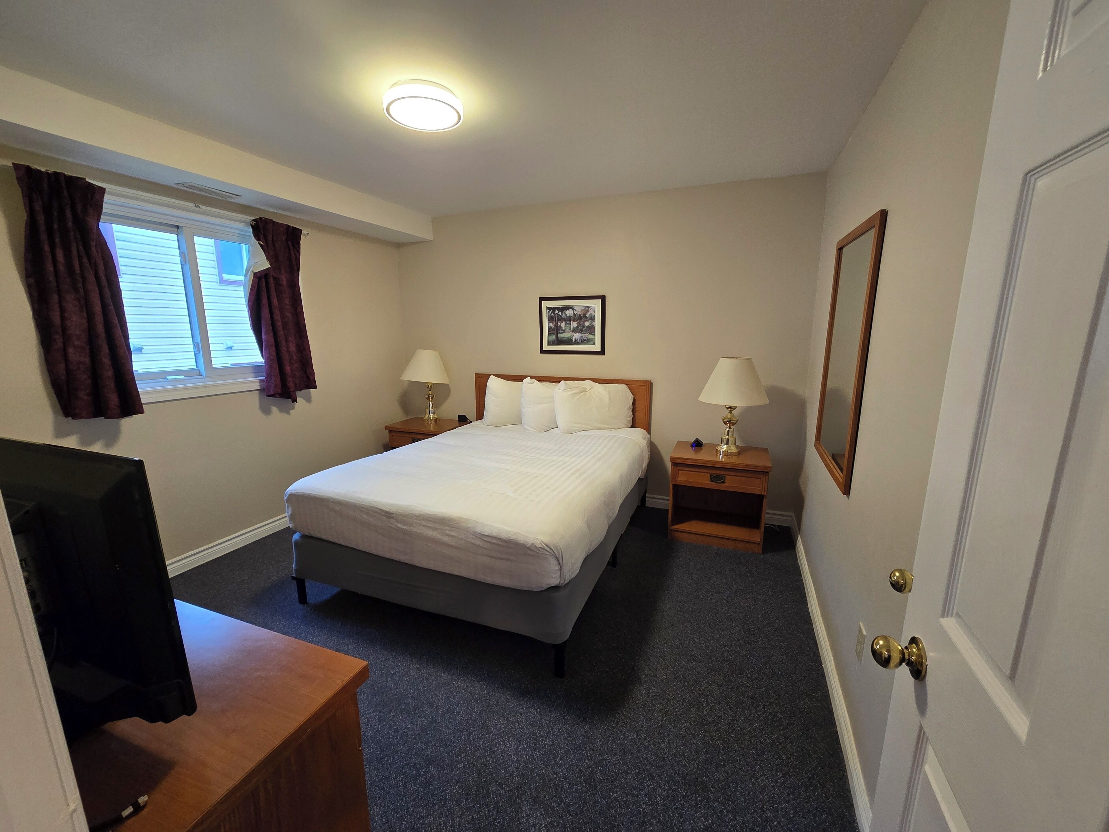A hotel room with a large bed, two bedside tables with lamps, a flat-screen TV, and a window with curtains.