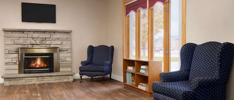 A living room with a stone fireplace, two patterned armchairs, a bookshelf, and a large window with a view of trees.