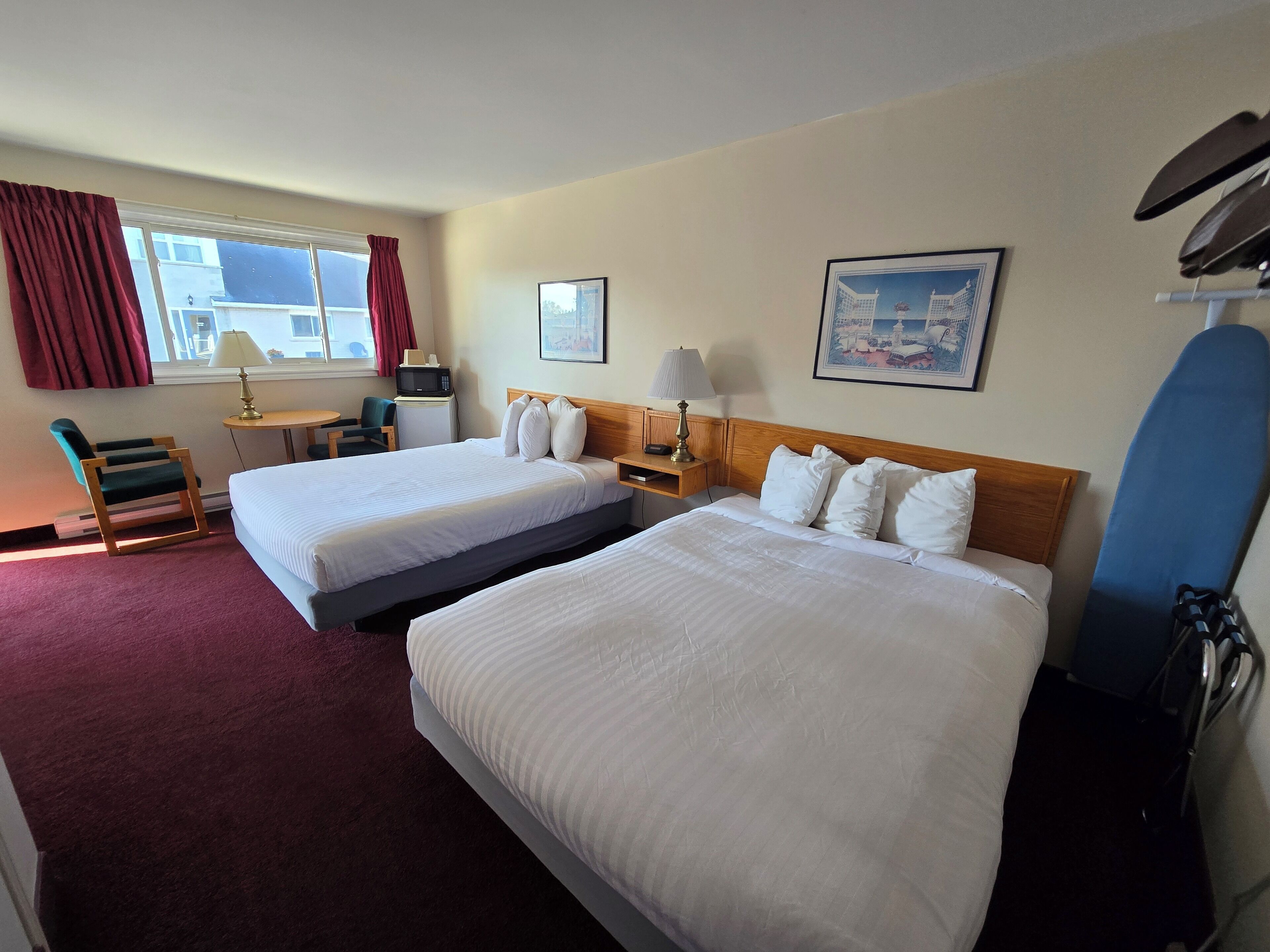 Habitación de hotel con dos camas, un escritorio, una silla, un microondas, una lámpara, un cuadro en la pared y una tabla de snowboard apoyada contra la pared.