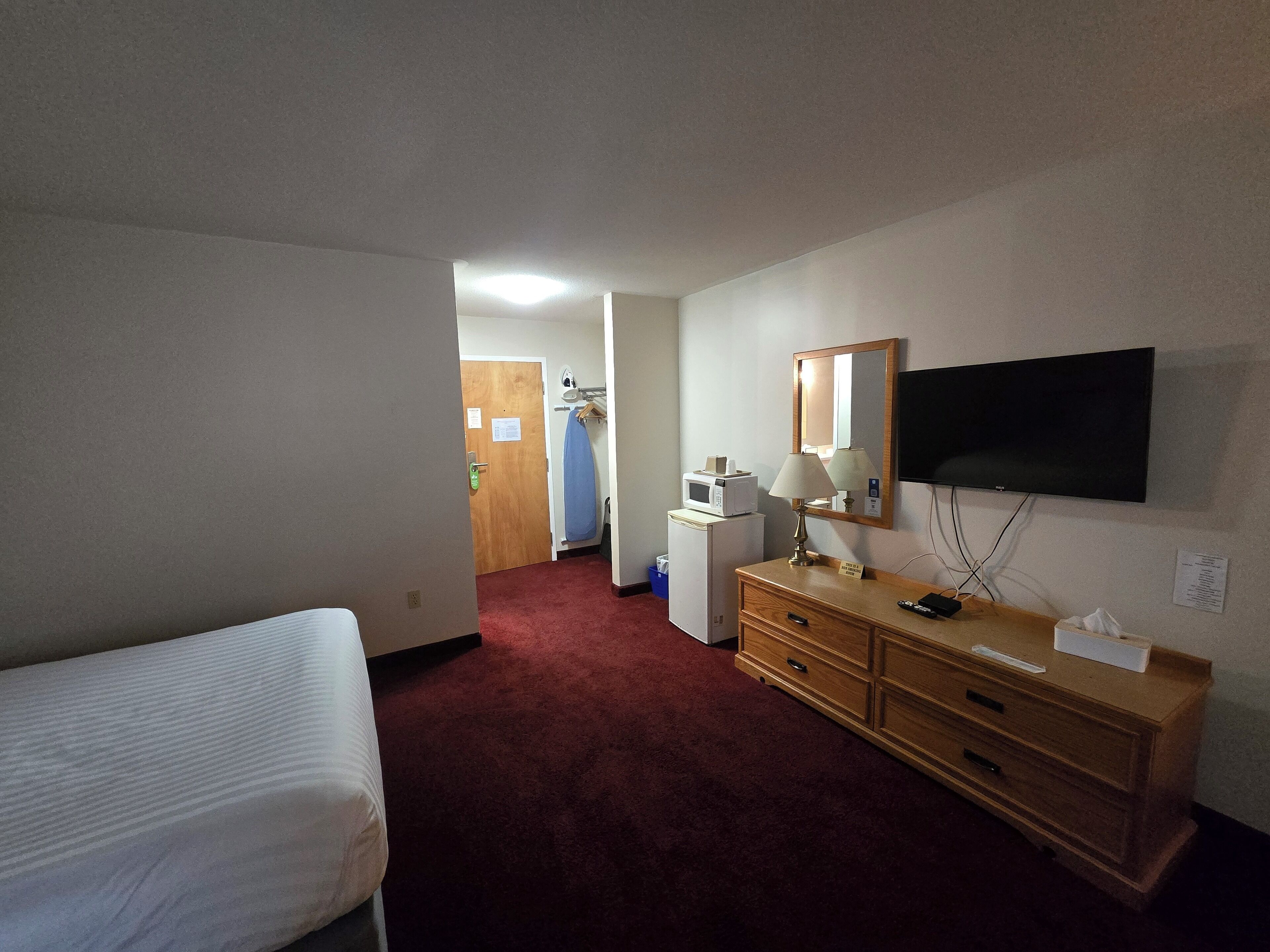 Een hotelkamer met een bed, een houten dressoir, een bureau met lamp, een magnetron, een televisie en een rode vloerbedekking.