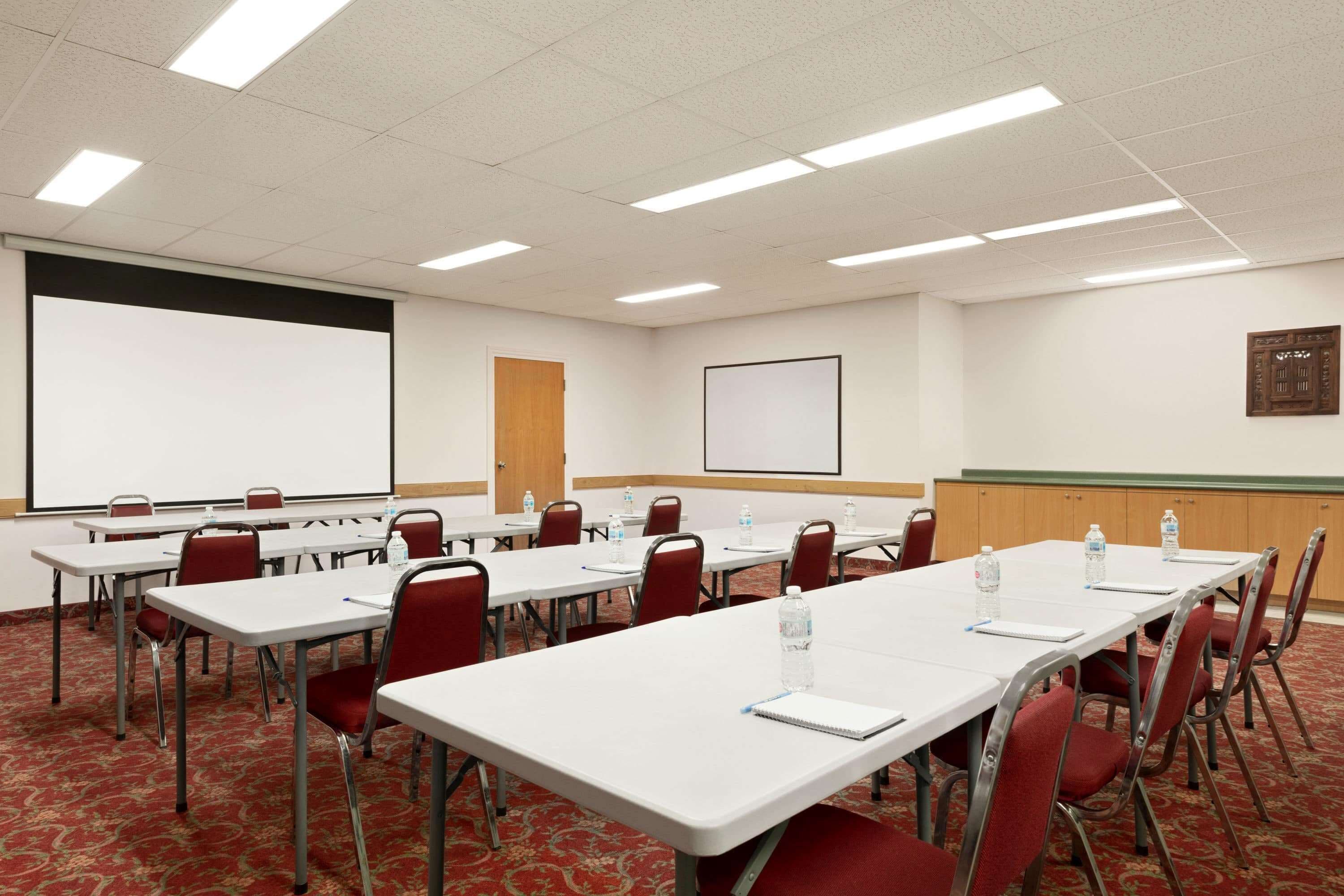 Un salón de conferencias con mesas blancas, sillas rojas y una pantalla de proyector.