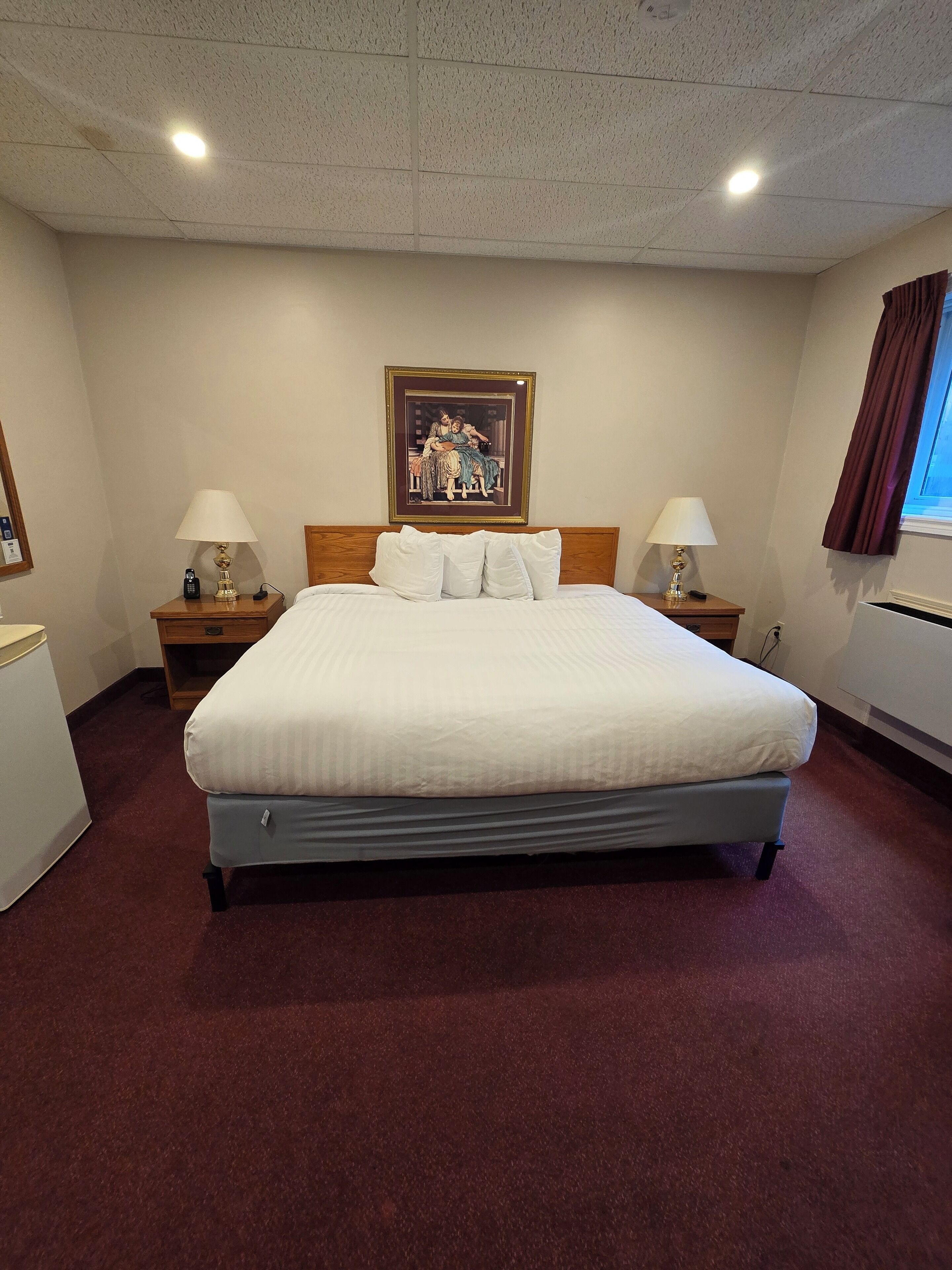 Quarto de hotel com uma cama grande, duas mesas de cabeceira com luminárias, um quadro na parede e uma janela com cortinas.