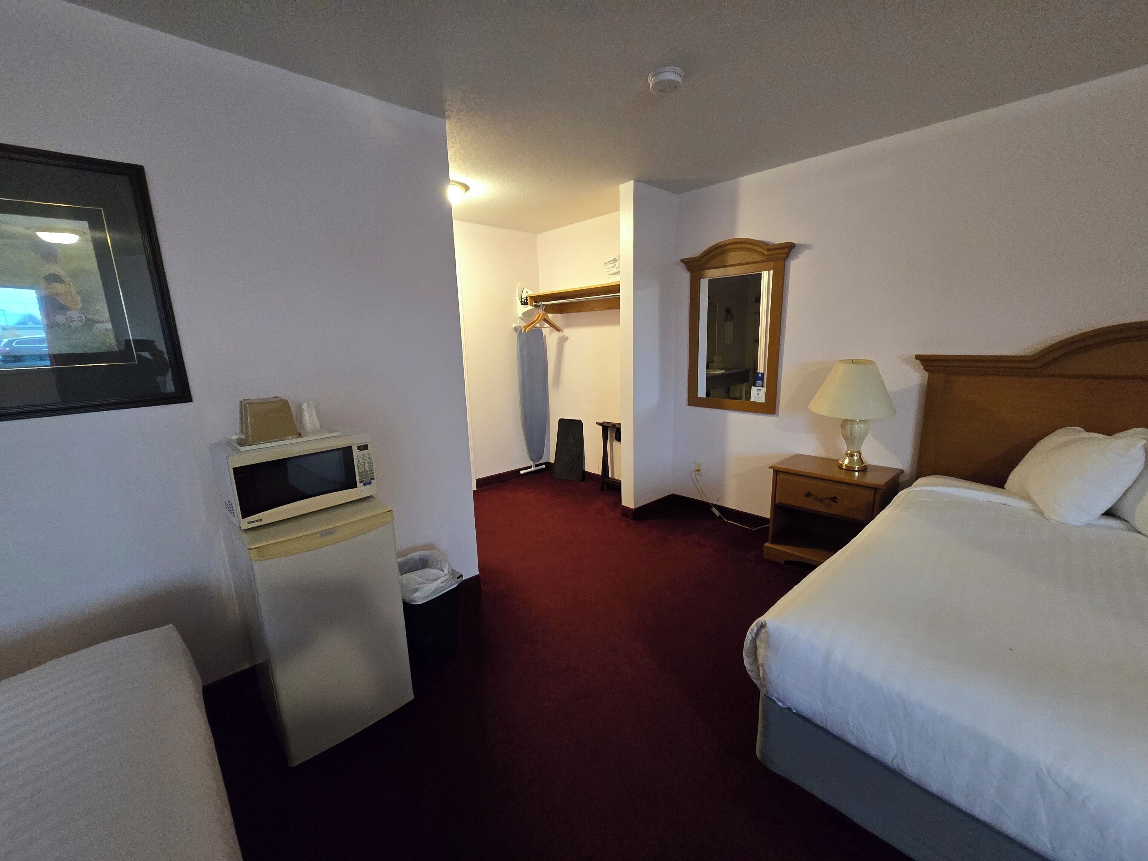 Une chambre d’hôtel comprenant un lit, une table de chevet avec une lampe, un miroir, un porte-manteau, un four à micro-ondes et une poubelle.