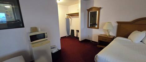 Una habitación de hotel con cama, una mesita de noche con lámpara, espejo, perchero, microondas y cubo de basura.