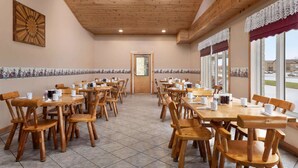 Une salle à manger spacieuse, avec des tables et des chaises en bois, un plafond en bois et de grandes fenêtres ornées de rideaux en dentelle.