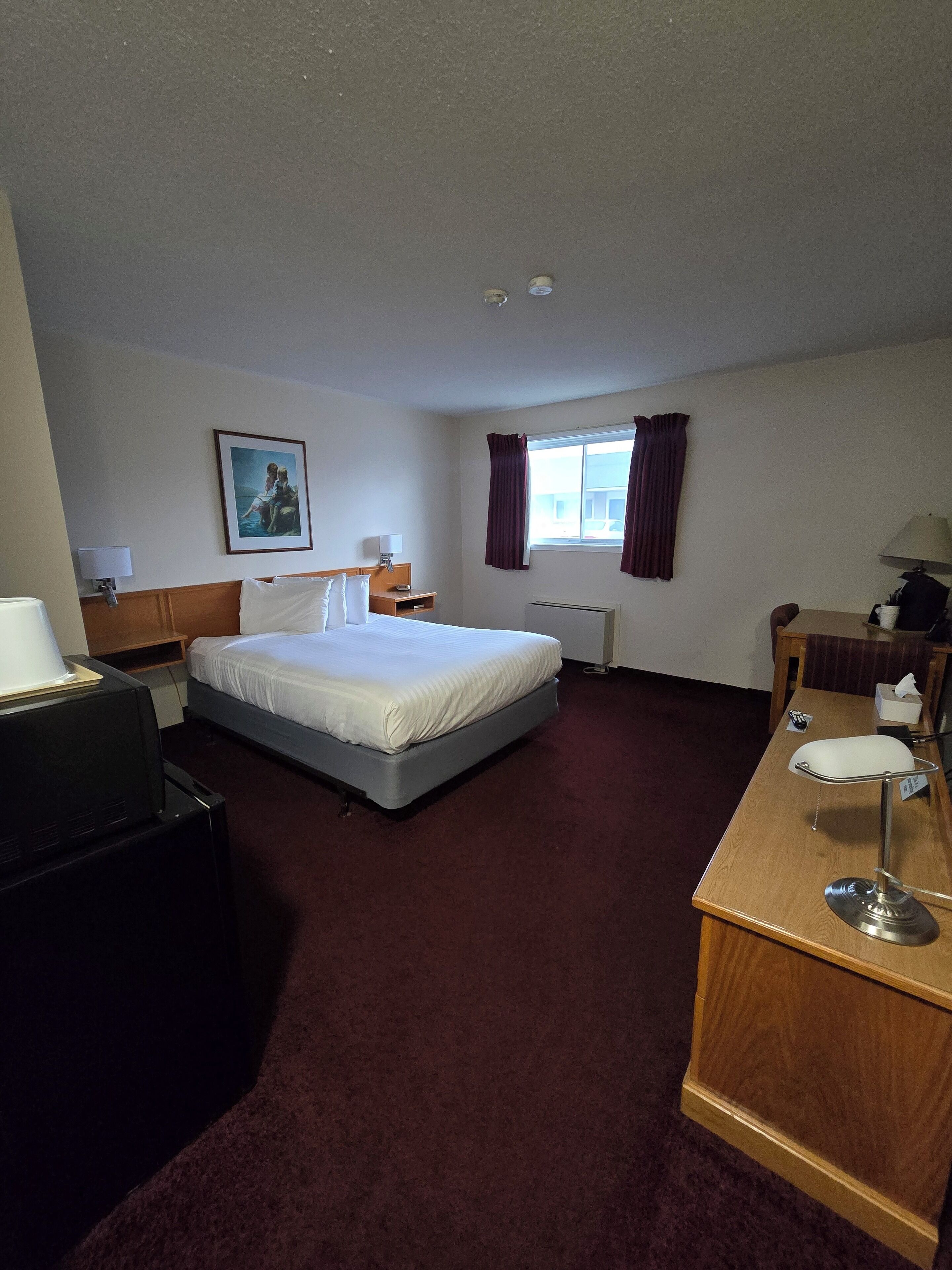 A hotel room with a single bed, a desk with a chair, a lamp, a window with curtains, and a picture on the wall.