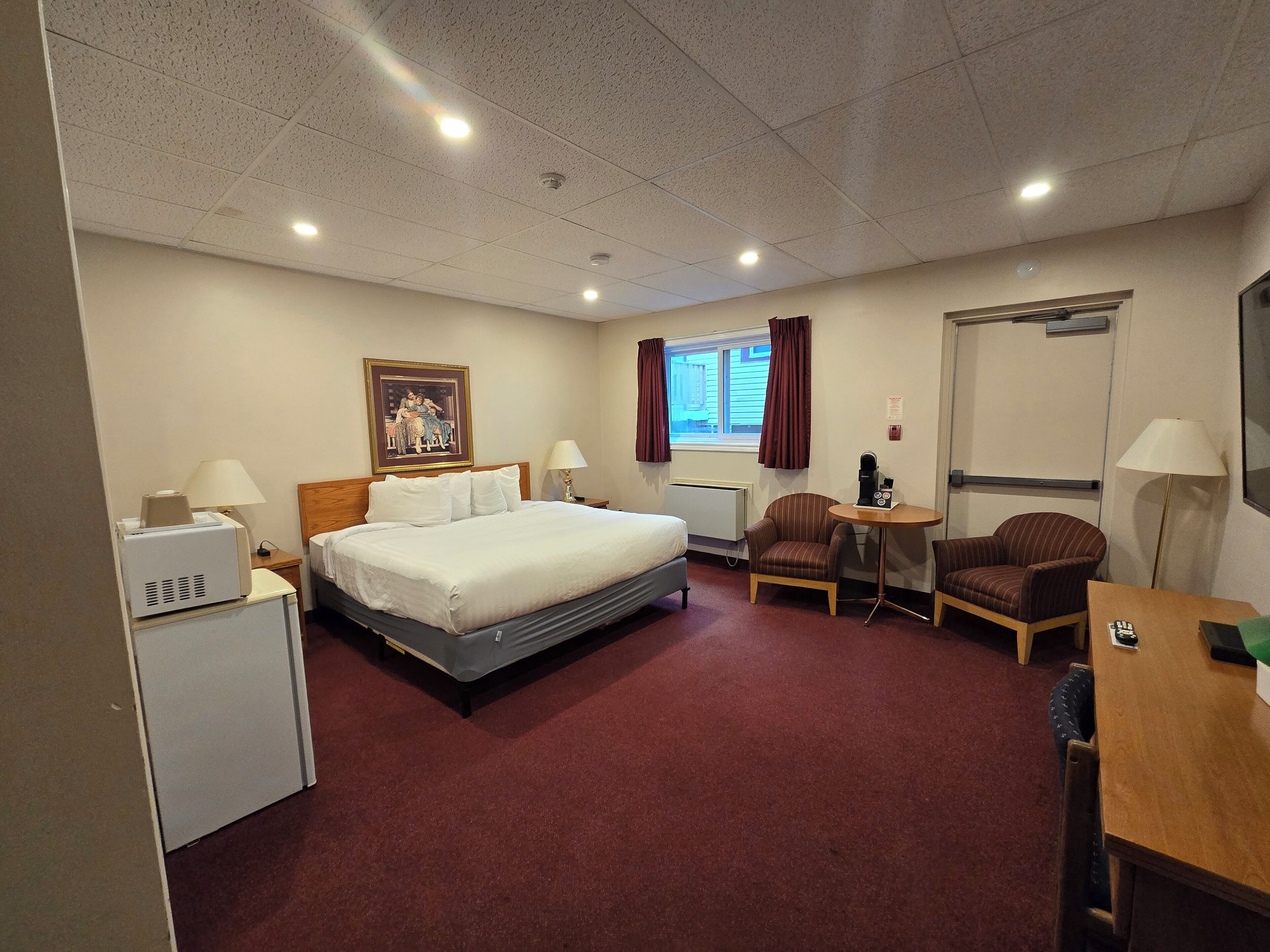 Une chambre d’hôtel avec un lit, un bureau, une chaise, une petite table, un radiateur, une fenêtre avec des rideaux et un tableau au mur.