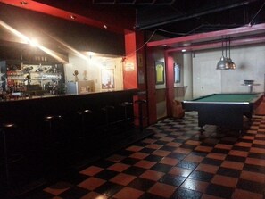 Un espace bar équipé d’une table de billard et de tabourets, doté d’un sol à carreaux et d’un comptoir où sont exposées des bouteilles.