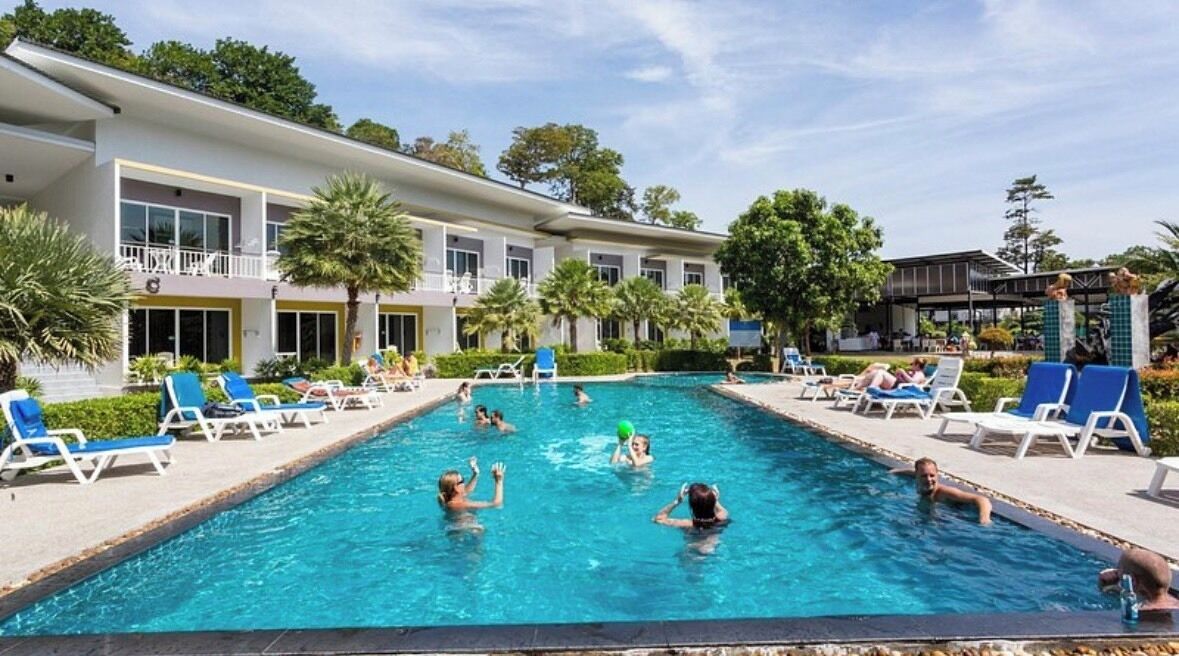 A swimming pool with people playing, surrounded by lounge chairs and a building with balconies.