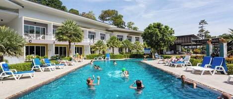 A swimming pool with people playing, surrounded by lounge chairs and a building with balconies.