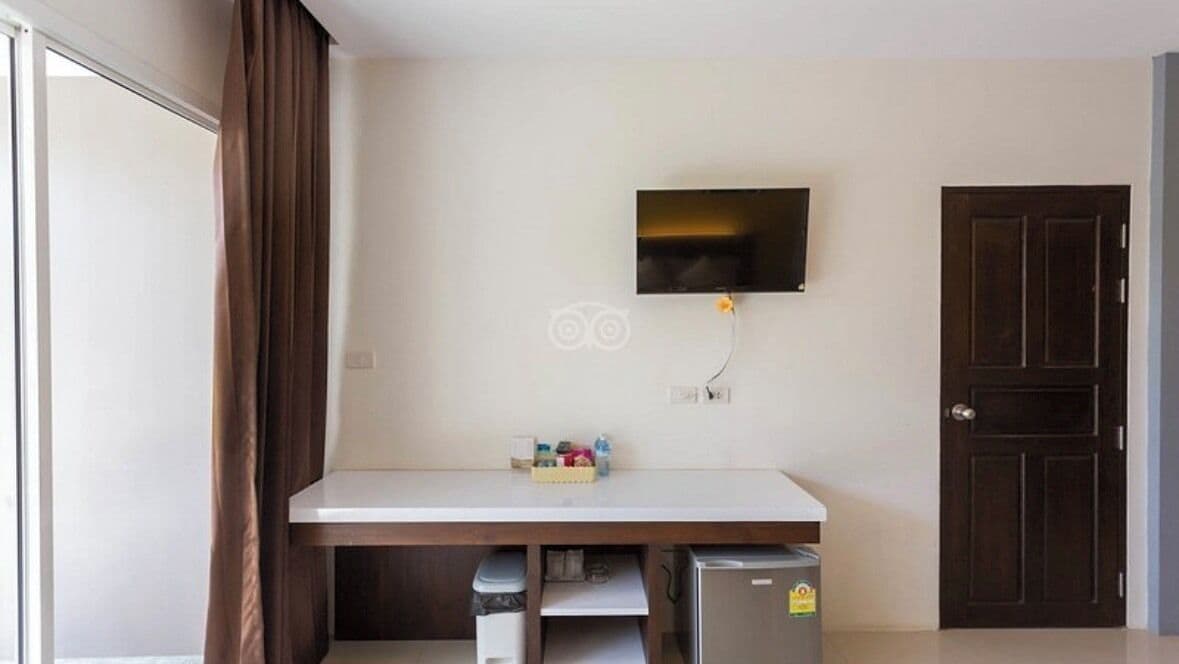 A room with a flat-screen TV mounted on the wall, a wooden desk with a trash bin underneath, and a dark wooden door.