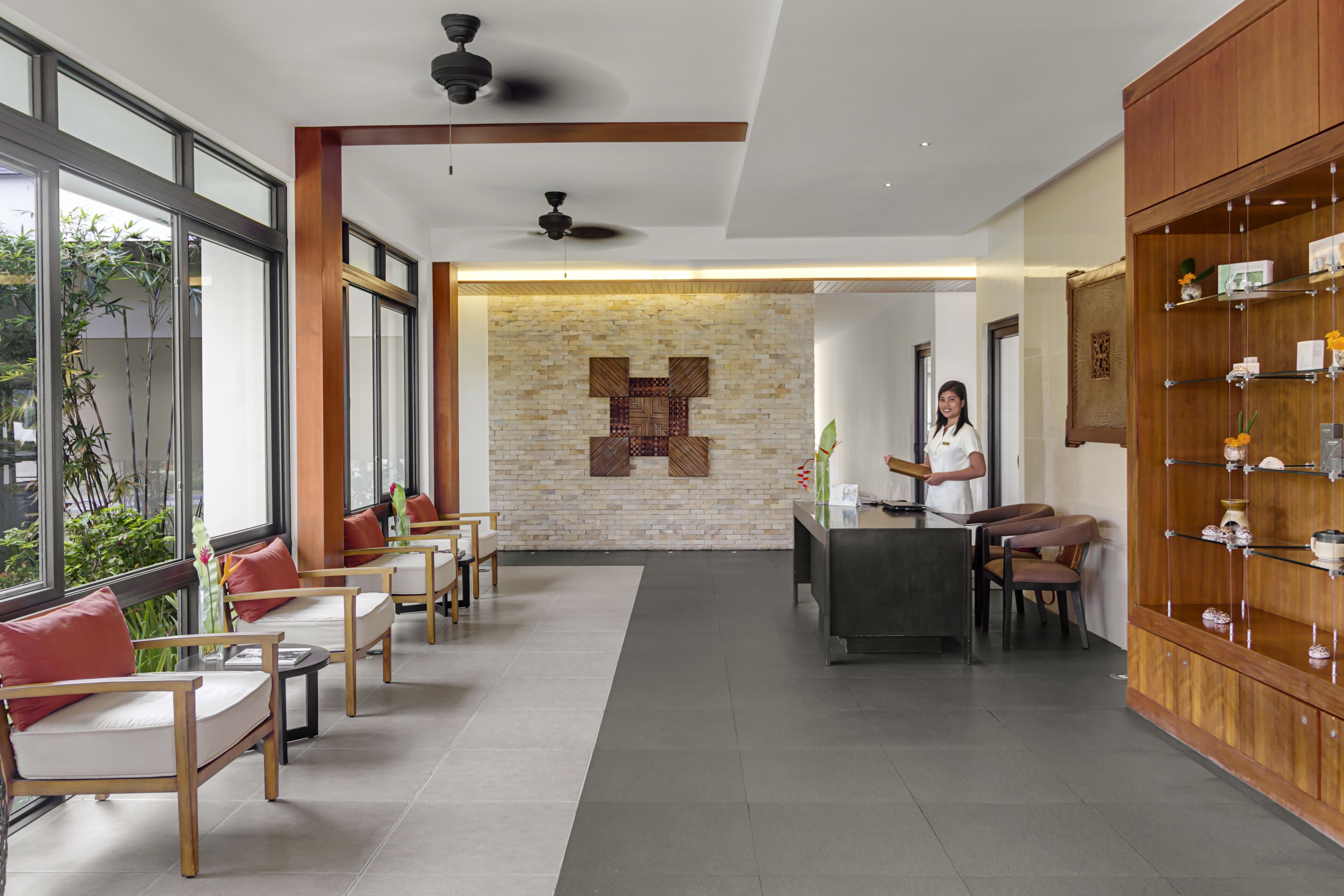 A modern hotel lobby with a reception desk, seating area with sofas and chairs, a wooden display cabinet, and large windows offering a view of greenery.
