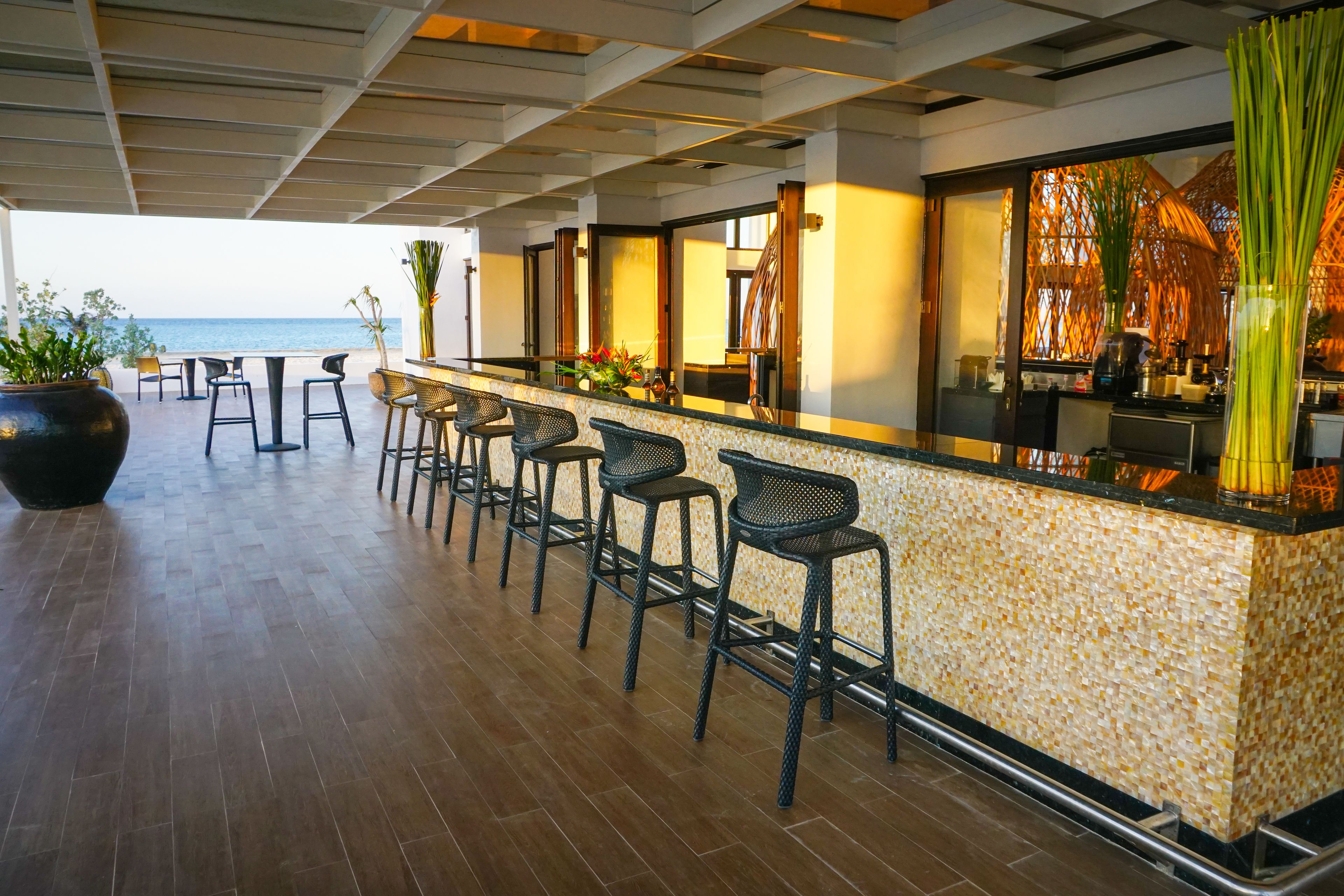 A bar area with a view of the sea, featuring a tiled floor, bar stools, and a counter with a patterned backsplash.