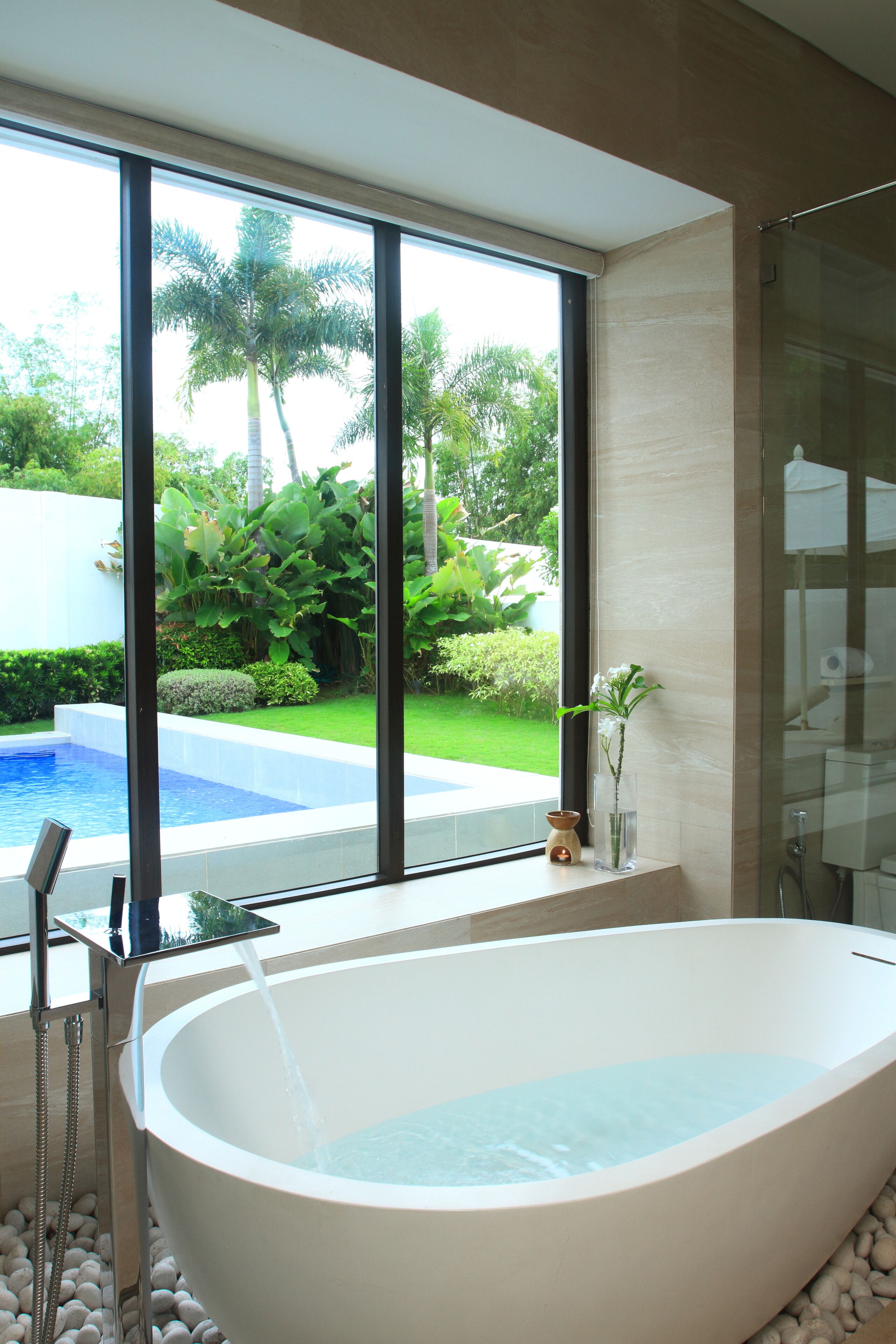 Un baño moderno con una bañera independiente, cabina de ducha con mampara de vidrio y vistas a una piscina y jardín.
