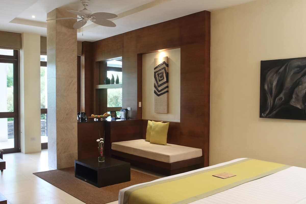 Une chambre d’hôtel moderne comprenant un lit, un coin salon avec un canapé et une table basse, une œuvre d’art accrochée au mur et une grande fenêtre offrant une vue sur l’extérieur.