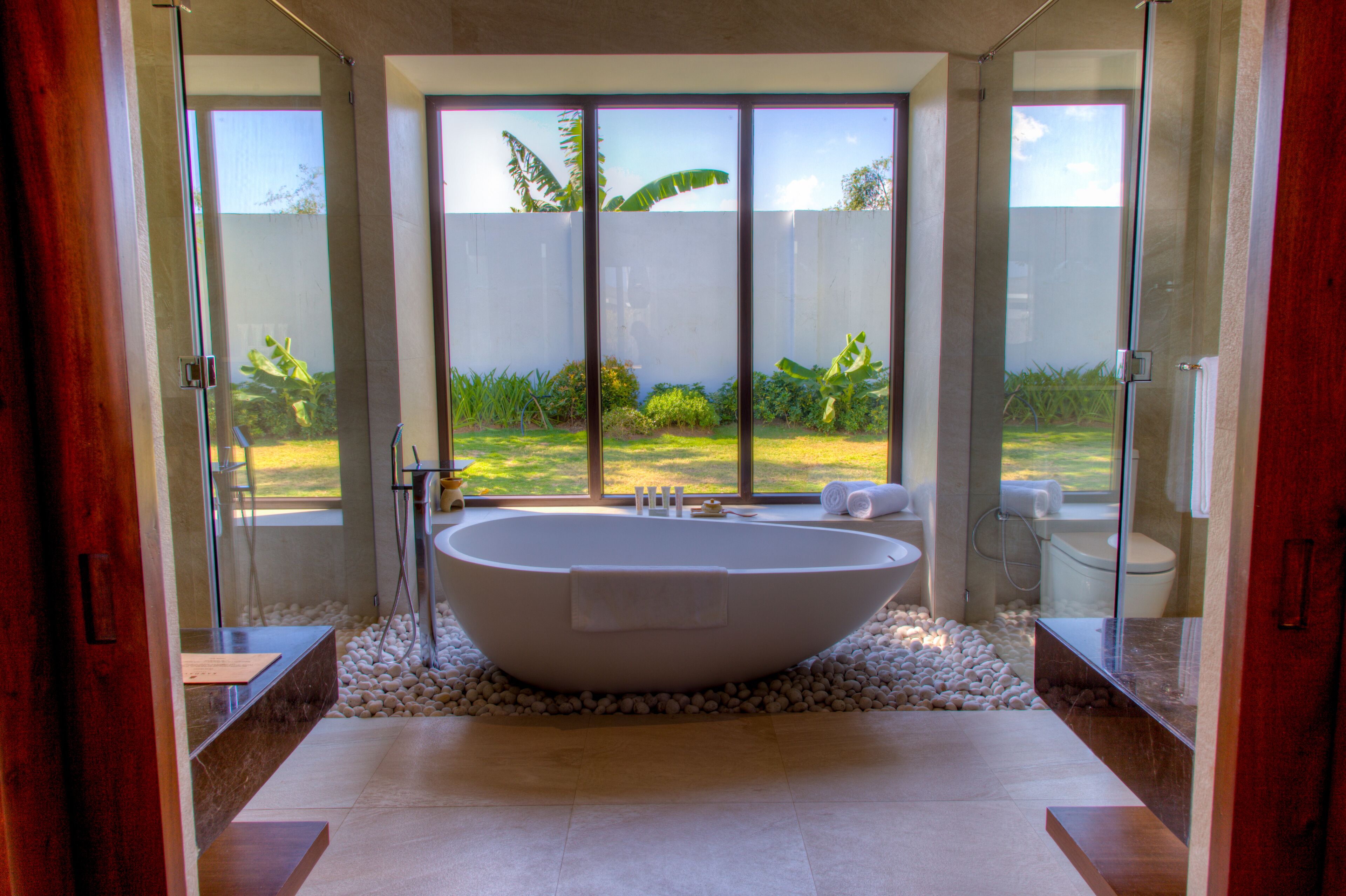 A modern bathroom with a freestanding bathtub, a glass shower enclosure, and a view of a lush garden through large windows.