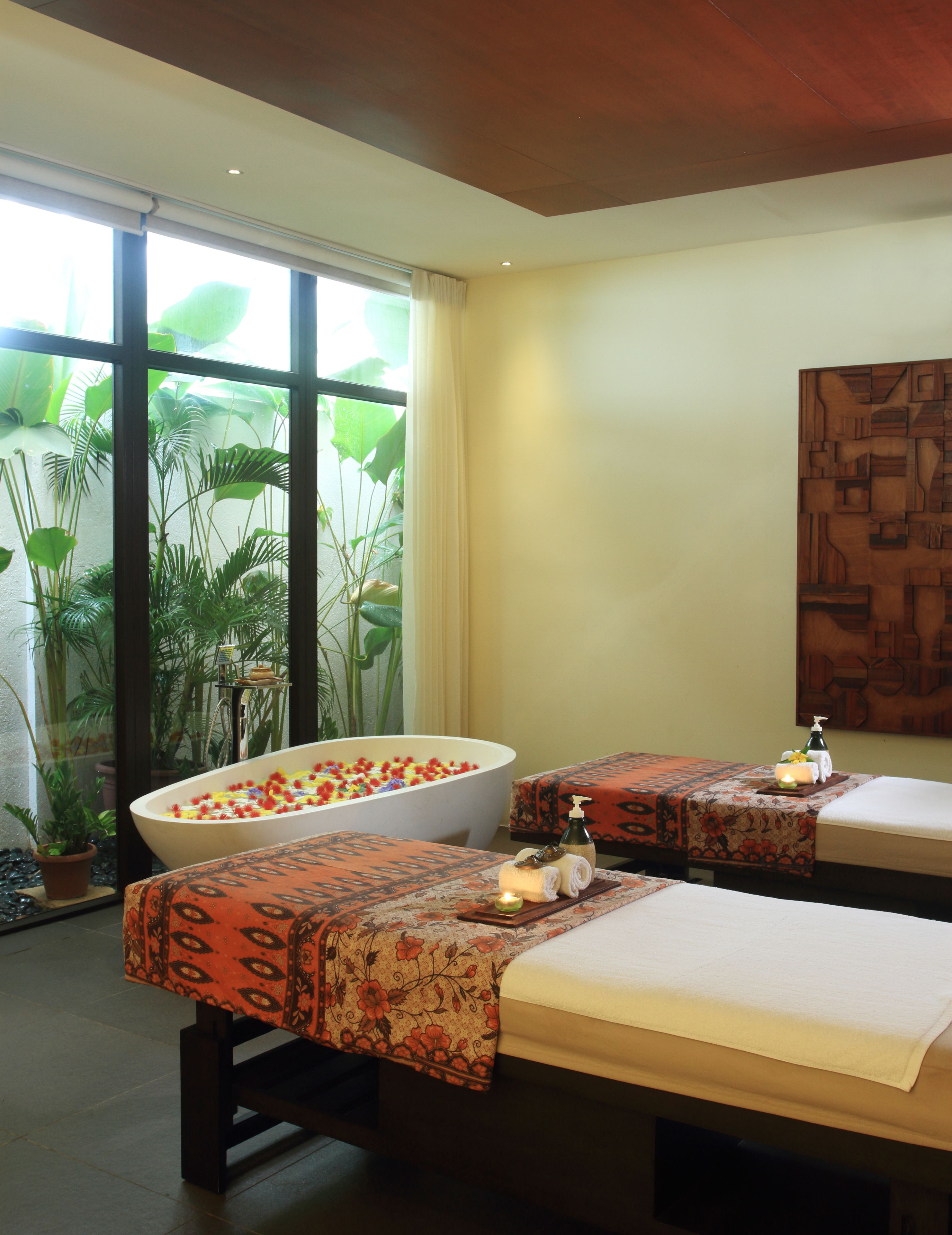 Ein Wellnessraum mit einer großen Badewanne, zwei Massageliegen und einem großen Fenster mit Pflanzen.