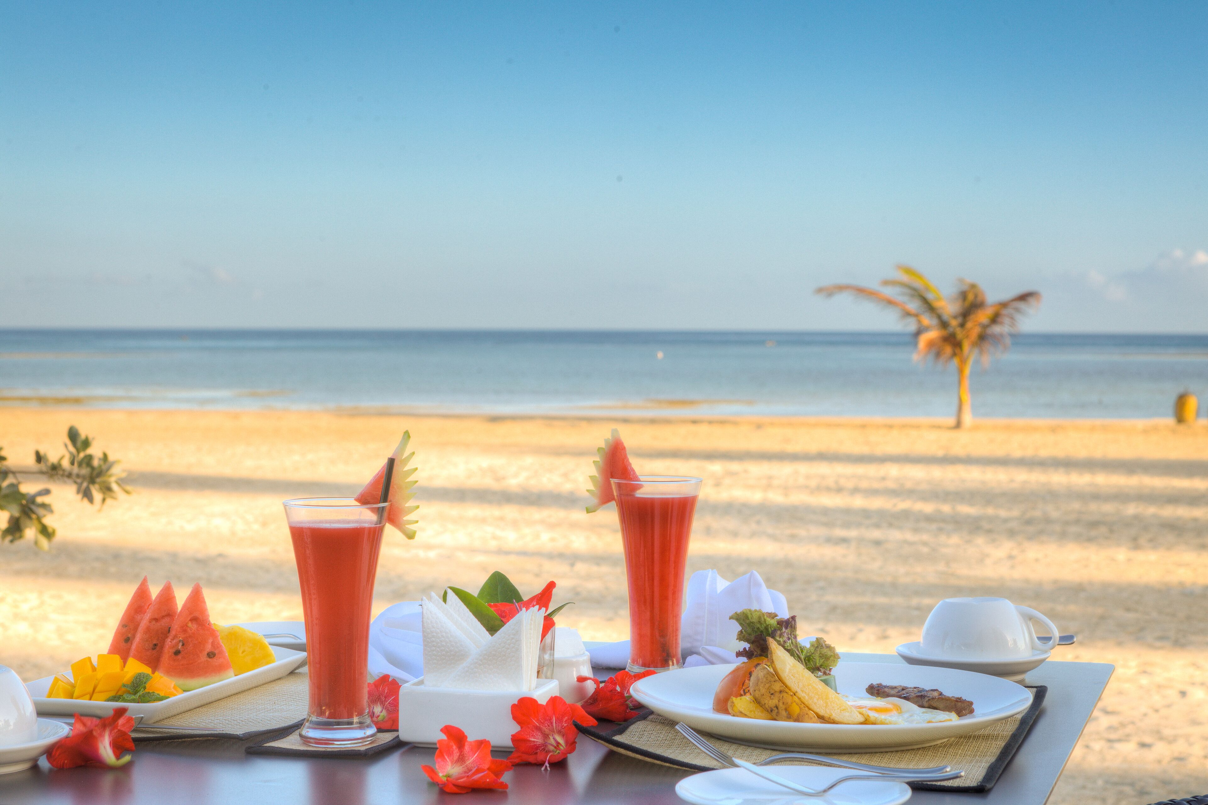 A table set with breakfast items including watermelon, orange slices, and a glass of red juice, with a beach and palm tree in the background.