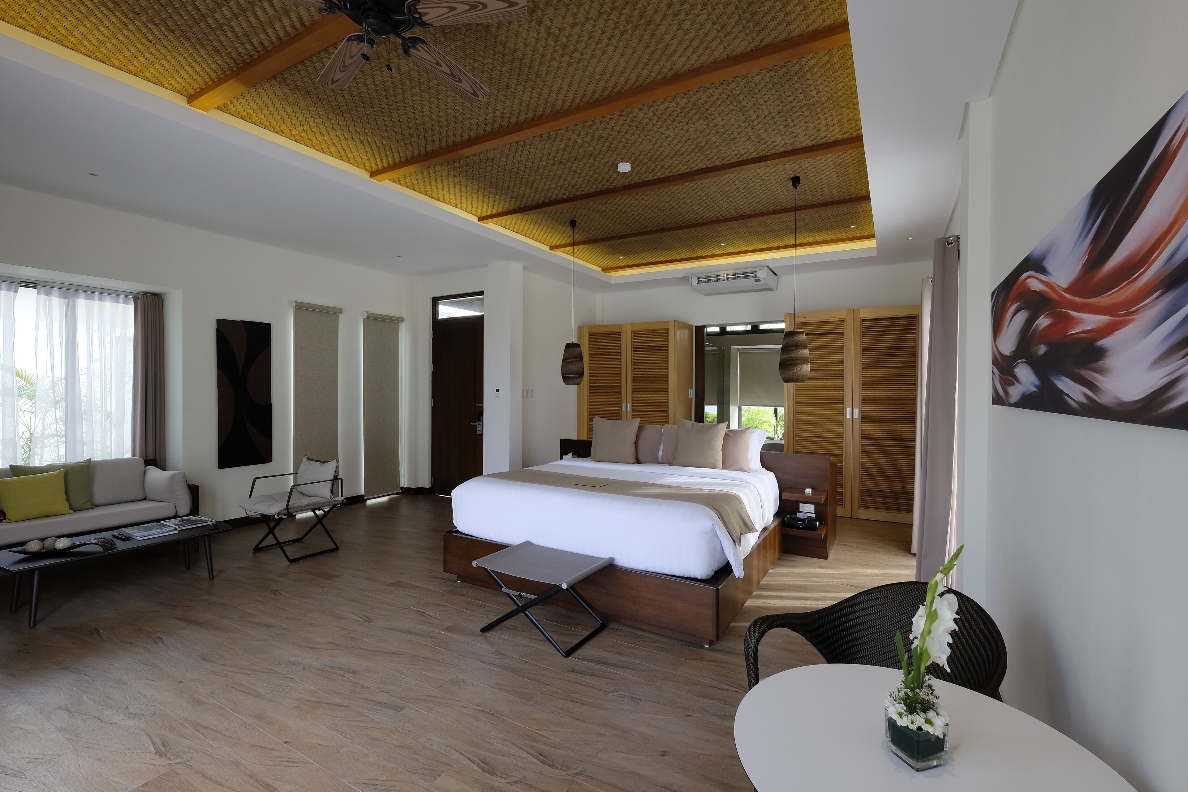 A modern hotel room with a large bed, a sitting area with a sofa and armchairs, a dining table with a vase of flowers, and a ceiling with wooden beams.