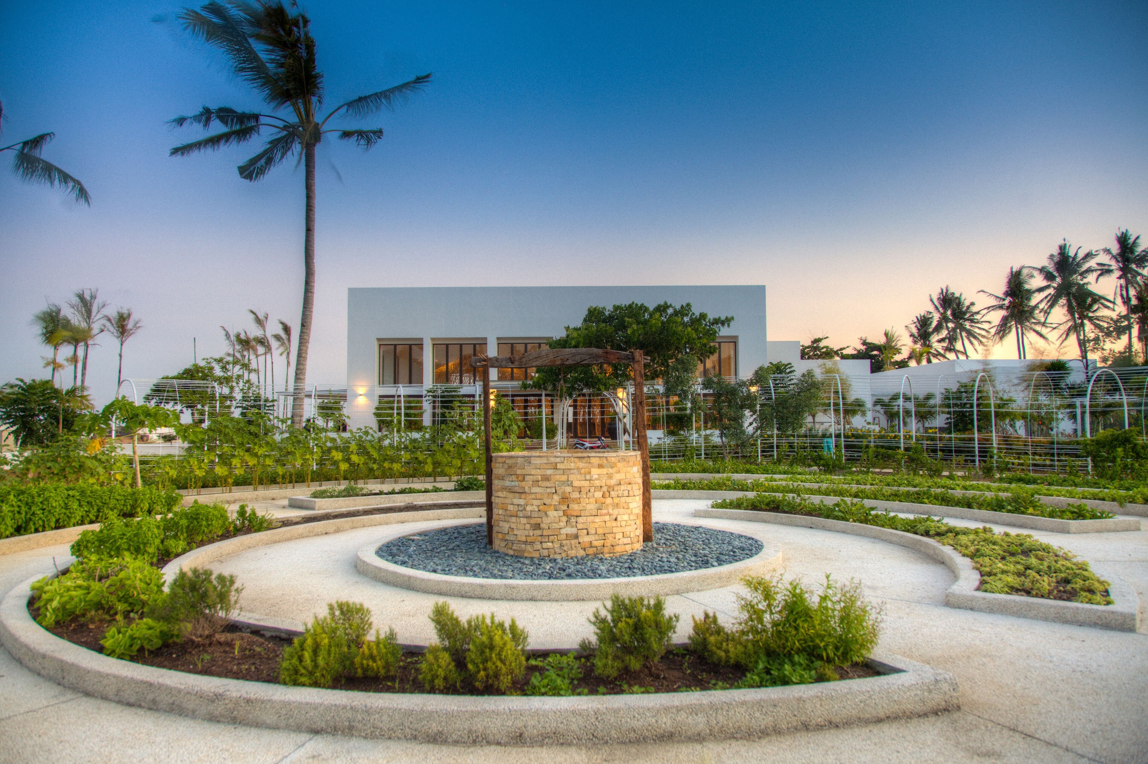 A landscaped garden with a central stone structure, palm trees, and a modern building in the background.